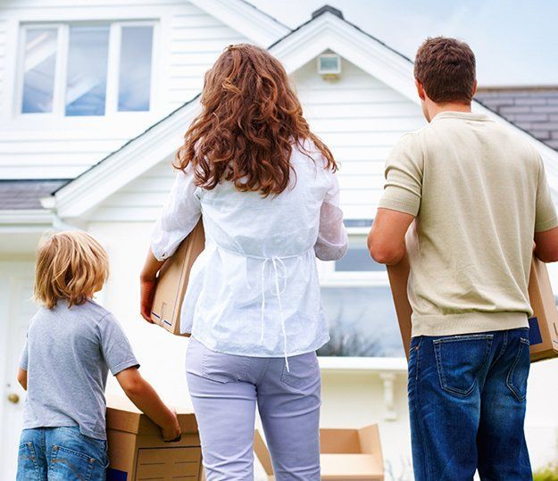 Une famille portant des cartons en direction d’une maison blanche, vue de dos