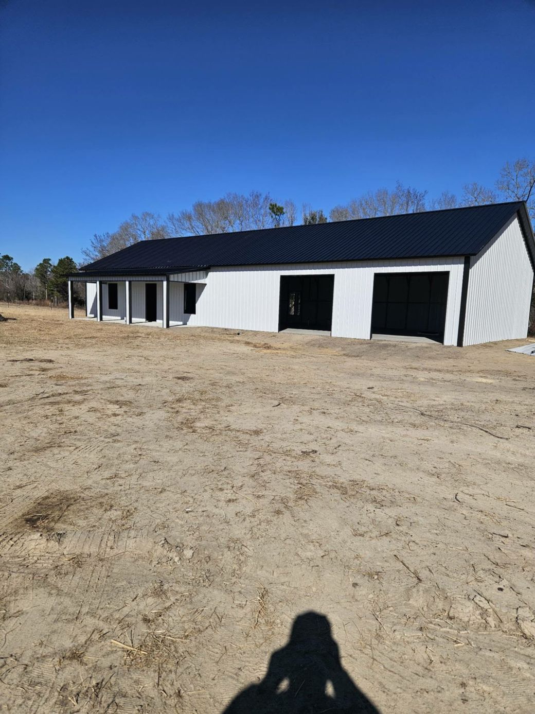 White barn-style building with black roof and doors on a bare dirt lot under a clear blue sky.
