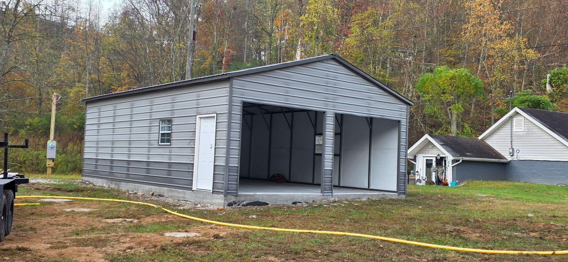 Gray metal shed under construction in a wooded yard, with open front doors and a small house nearby