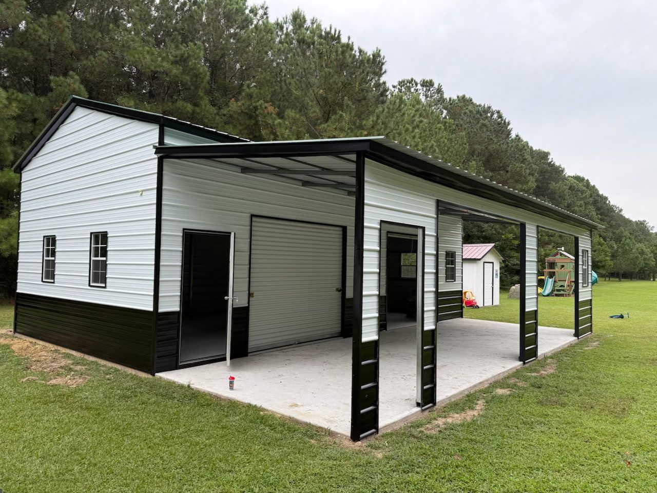 White metal outbuilding with covered porch, open garage bay, and black trim on a grassy lot