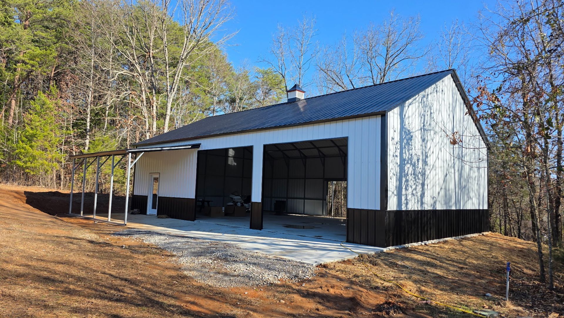 White metal barn with open garage bays on a wooded hillside under clear blue sky