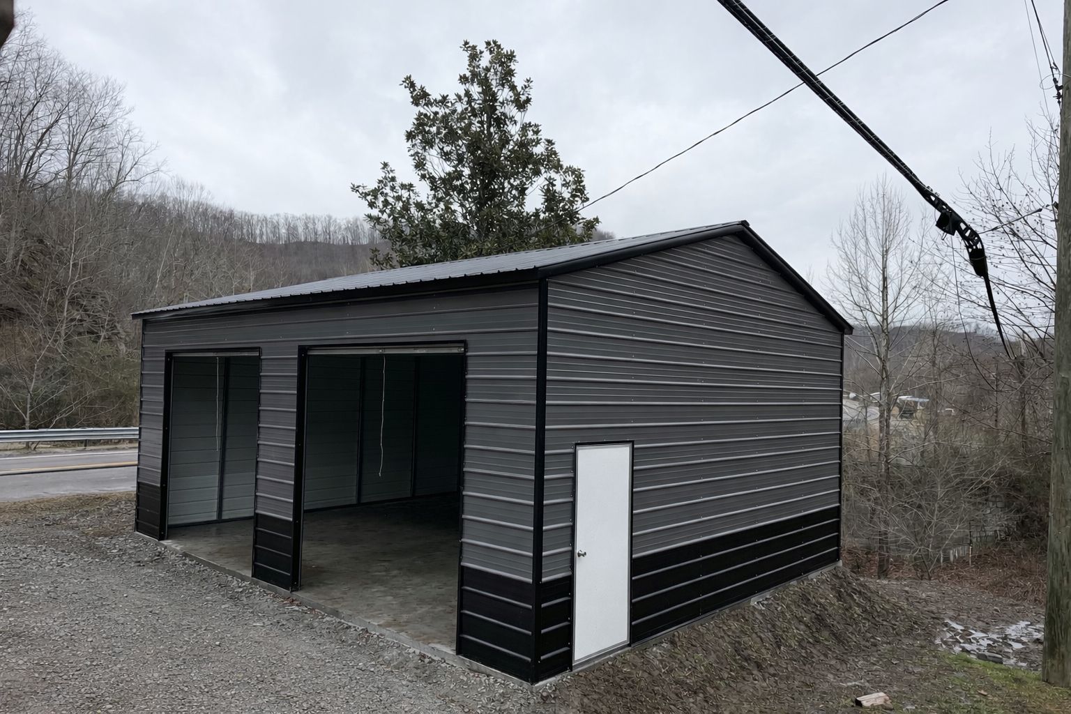 Dark gray shed-like building with open doorway on gravel, set beside a roadside in a wooded area