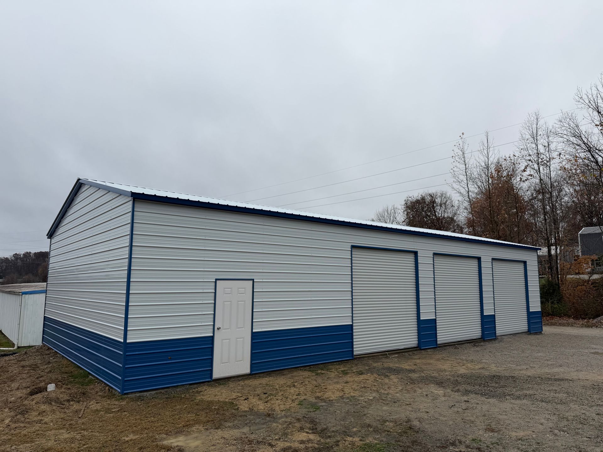 White-and-blue industrial building with large garage doors on a gravel lot under an overcast sky