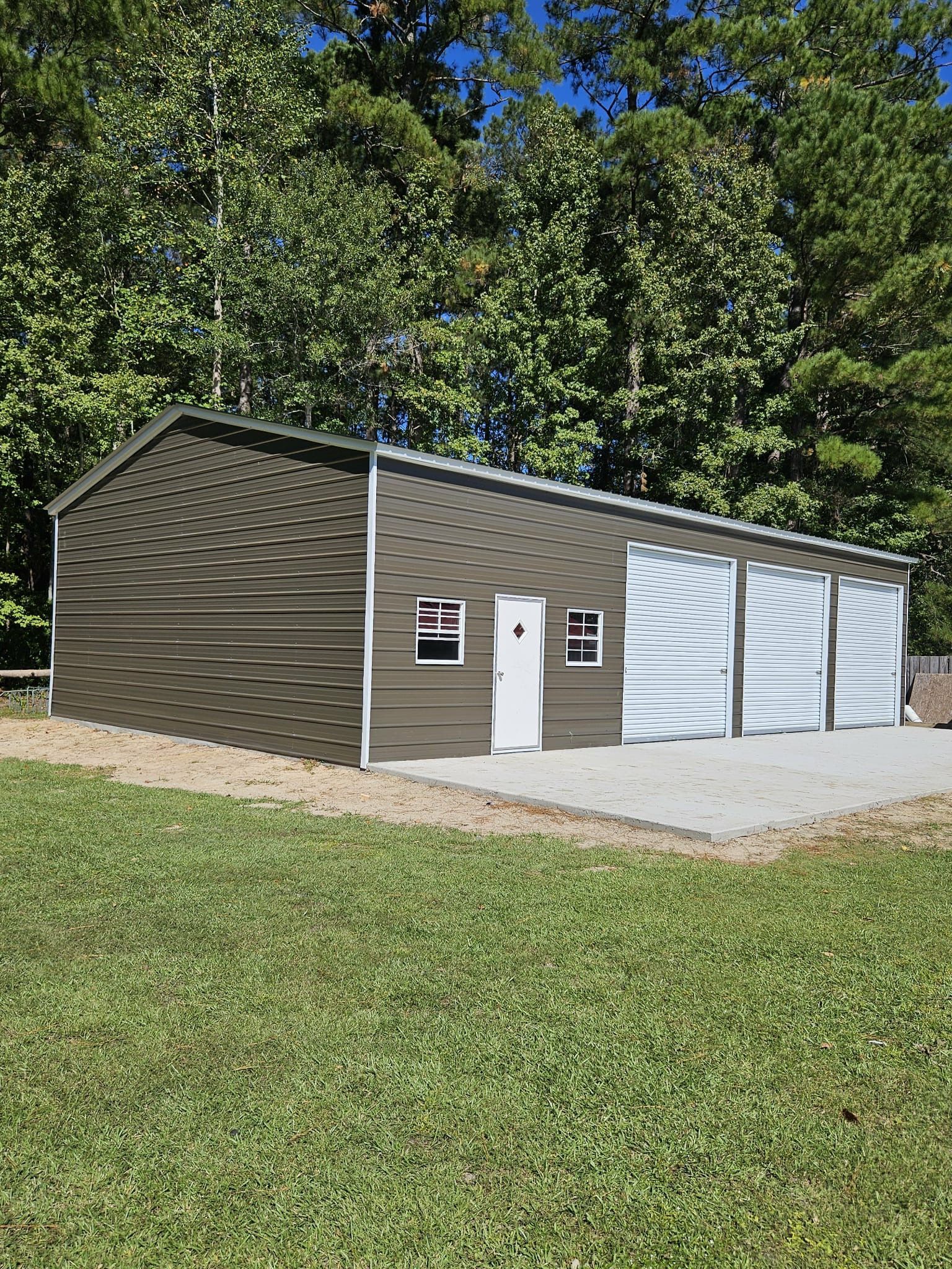 Small brown garage-style building with white doors and a paved driveway, surrounded by grass and trees