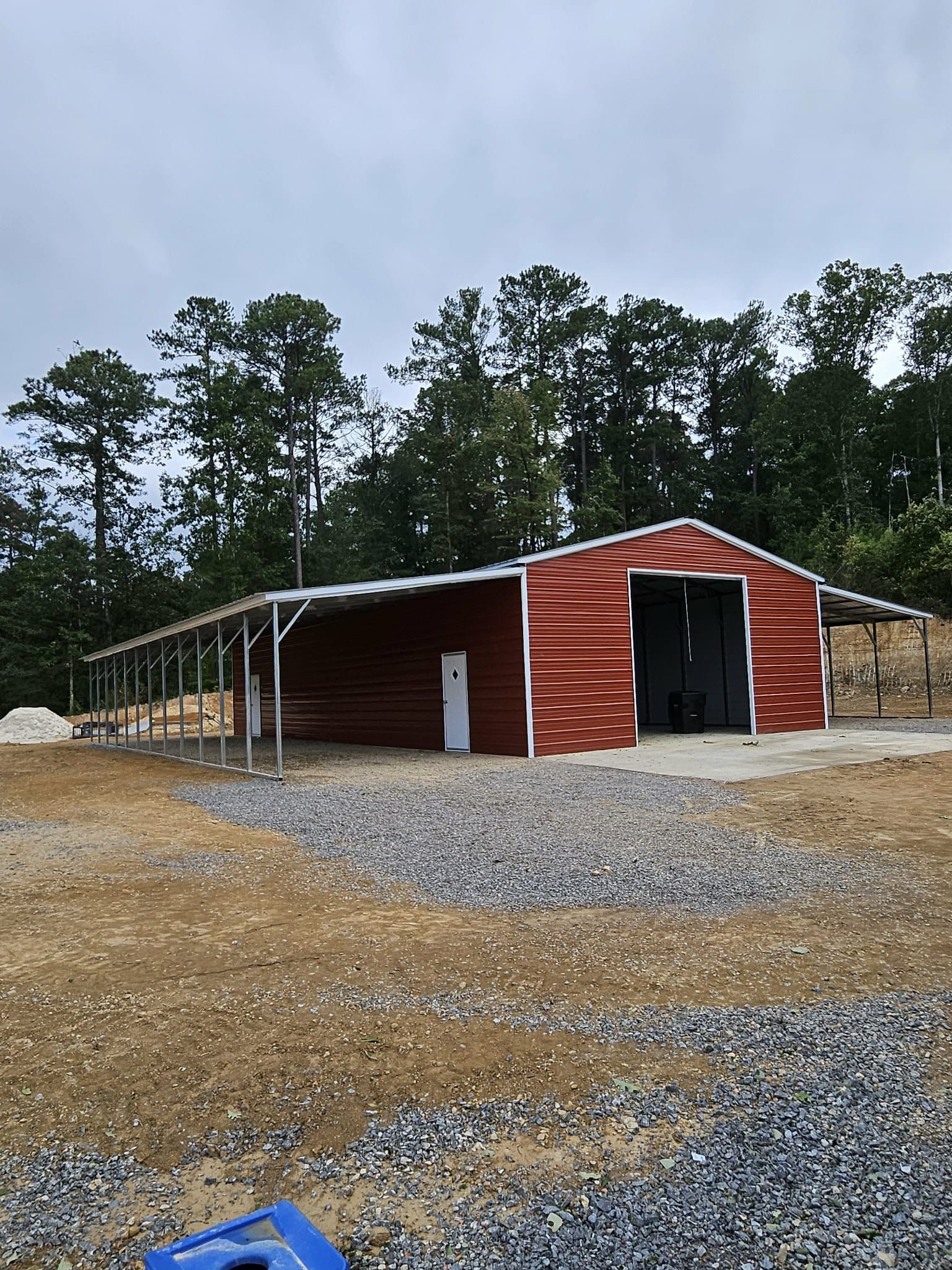 Red barn-style shed with open bays on a gravel lot, surrounded by trees under a cloudy sky
