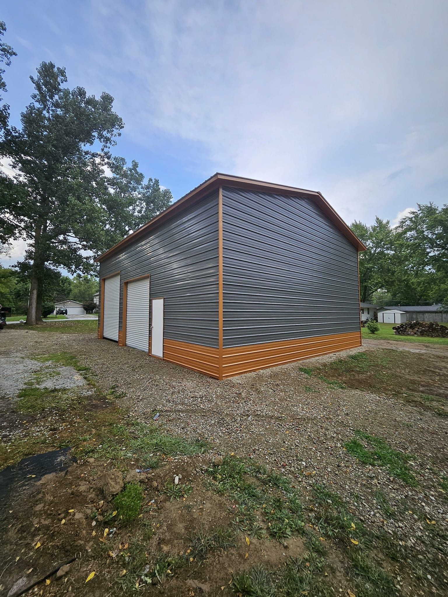 Small gray-and-orange patterned building with a white door, set on a gravel lot beside trees and grass.