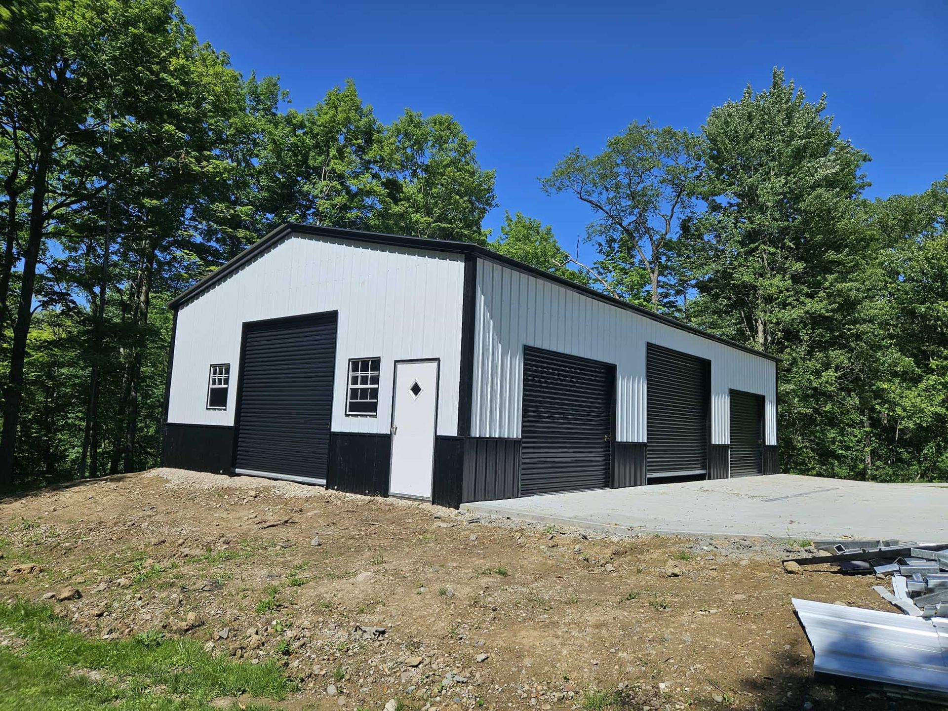 White metal barn-style building with black trim and large garage doors on a wooded lot