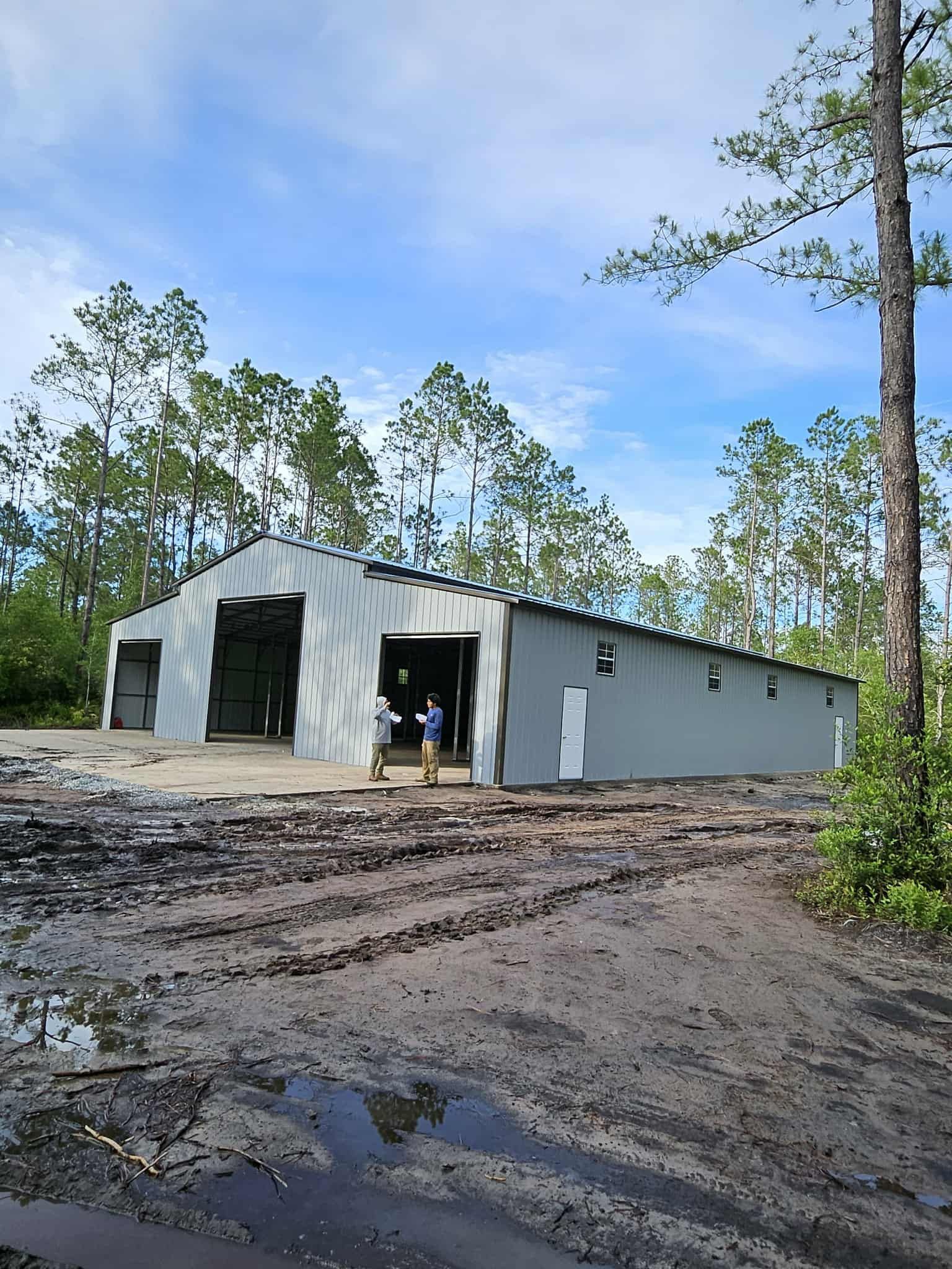 Gray metal warehouse with open doors on a muddy lot, trees and blue sky in the background.