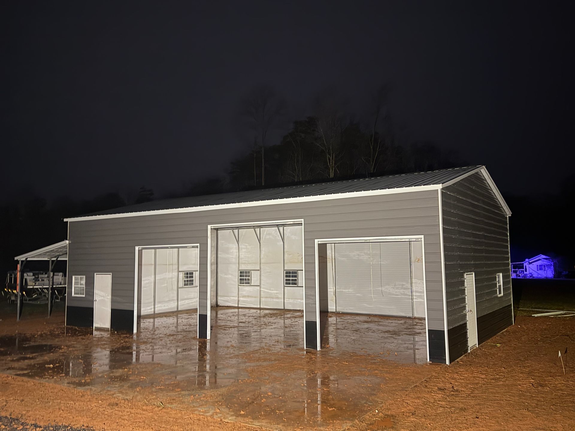 Gray metal building at night, lit by bright exterior lights, with open garage bays on a muddy lot.
