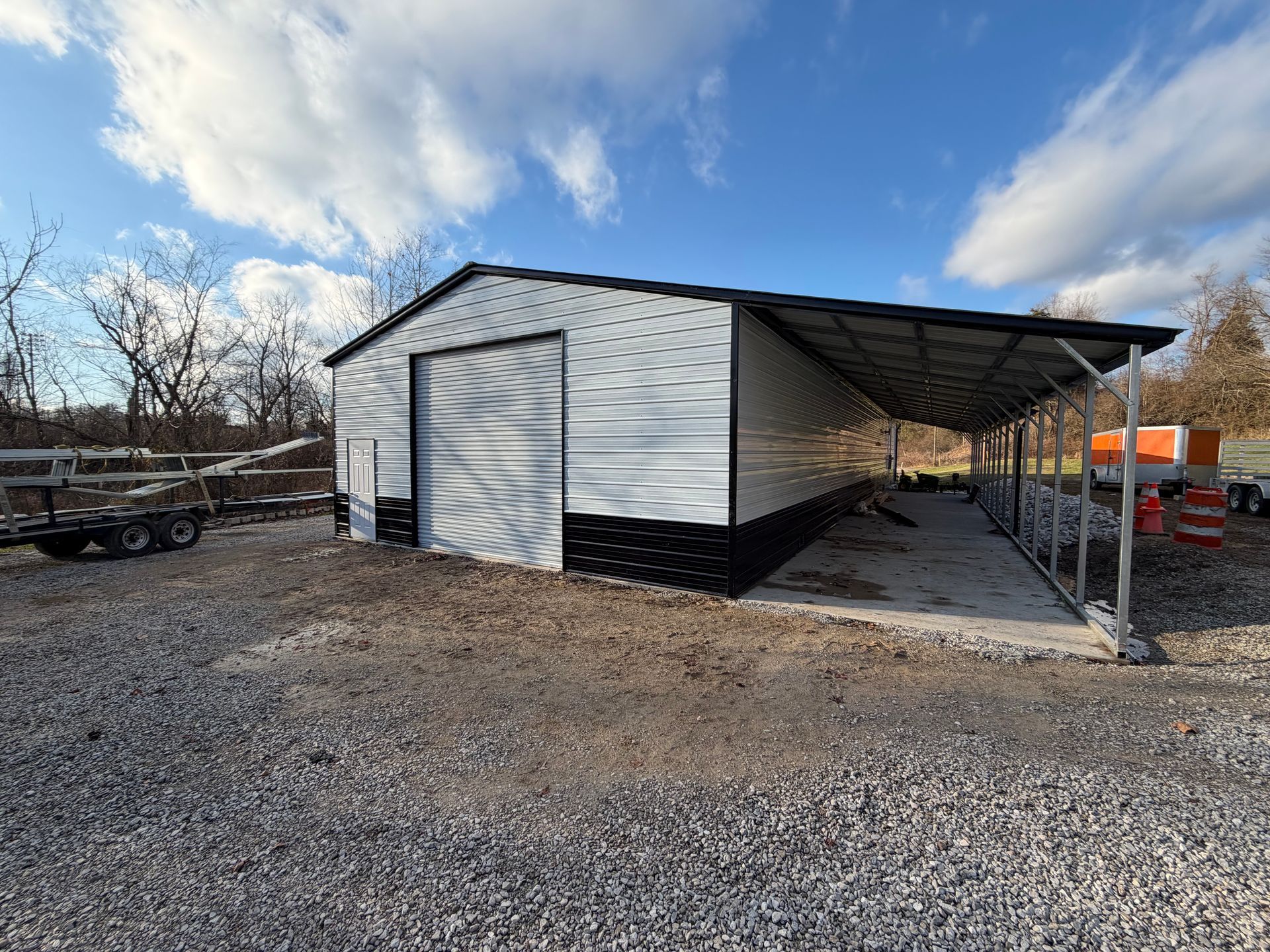 White metal shed with a large garage door and attached carport on a gravel lot under a blue sky.