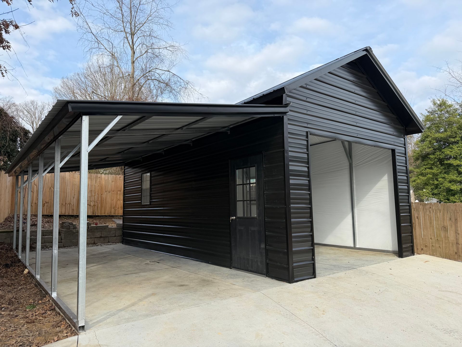 Black shed with covered carport and open garage-style door on a concrete driveway.