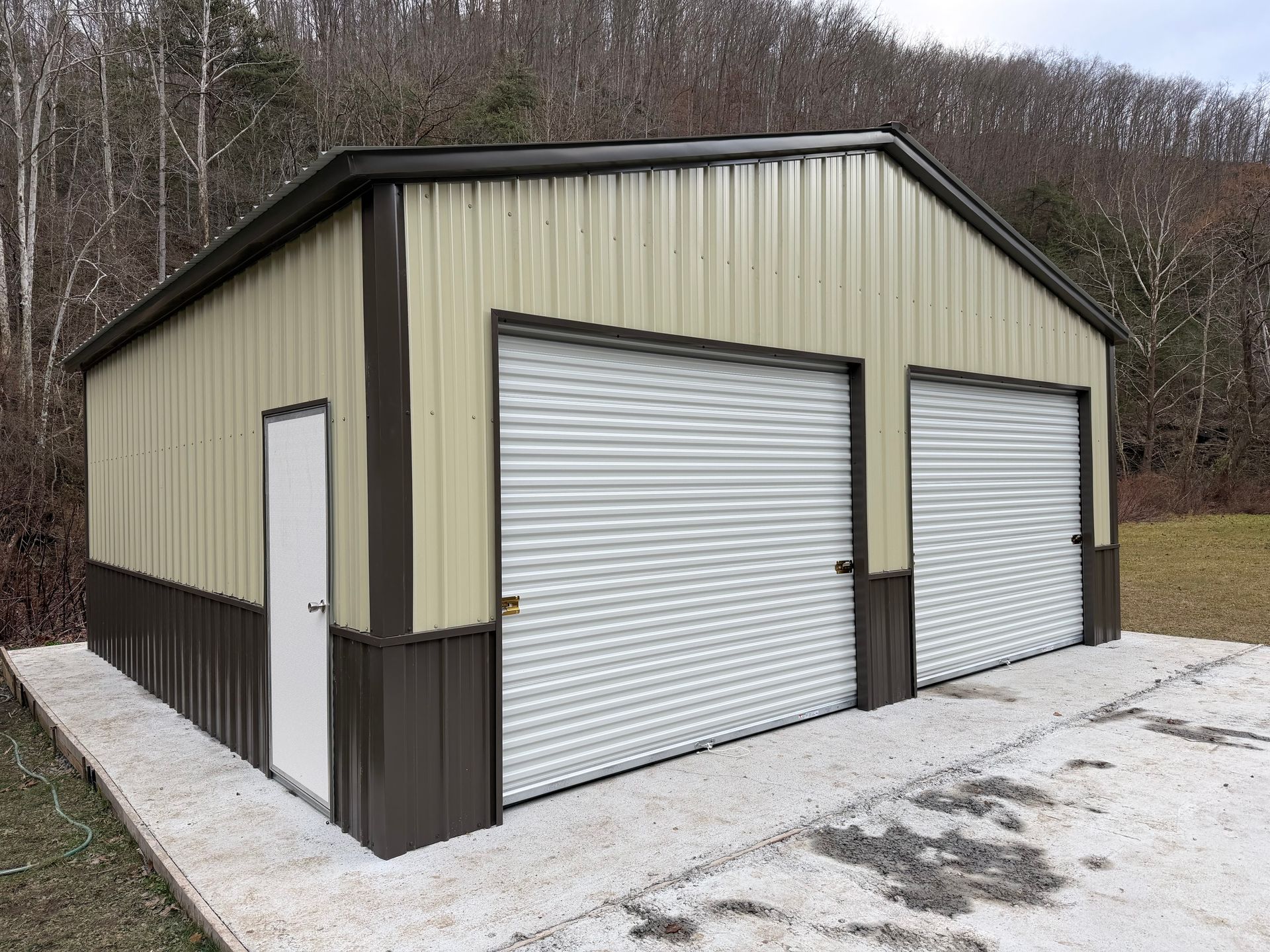 Tan metal garage with two roll-up doors and a side entry door on a gravel lot by trees