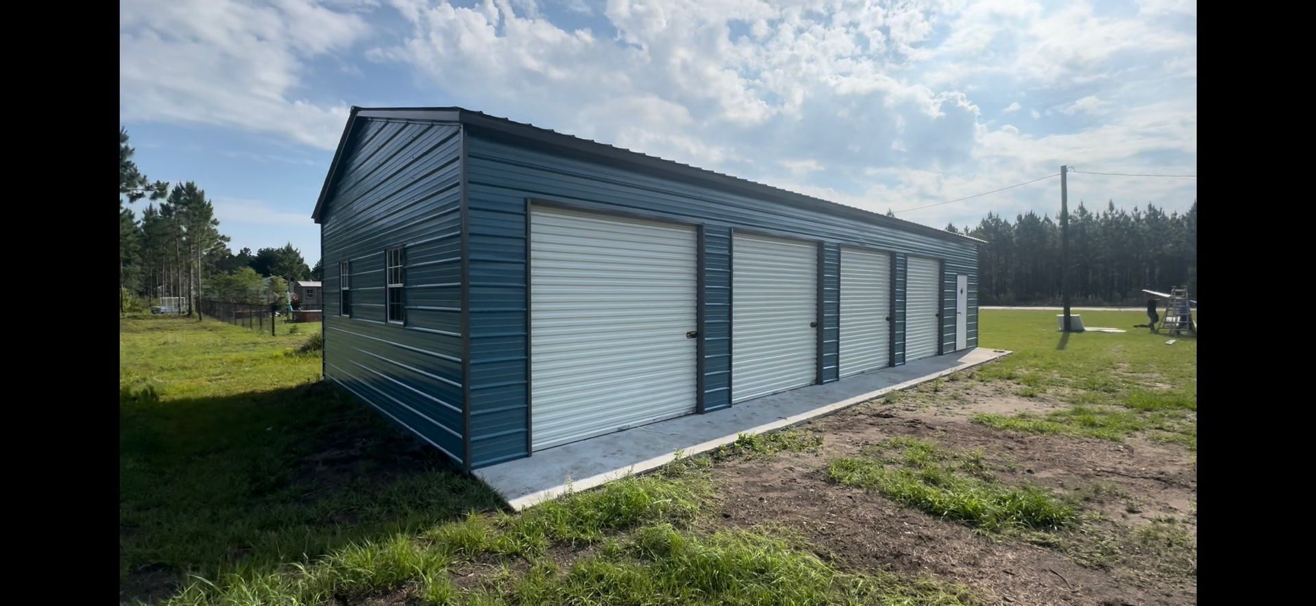 Blue metal storage building with multiple roll-up doors on a grassy lot under a partly cloudy sky