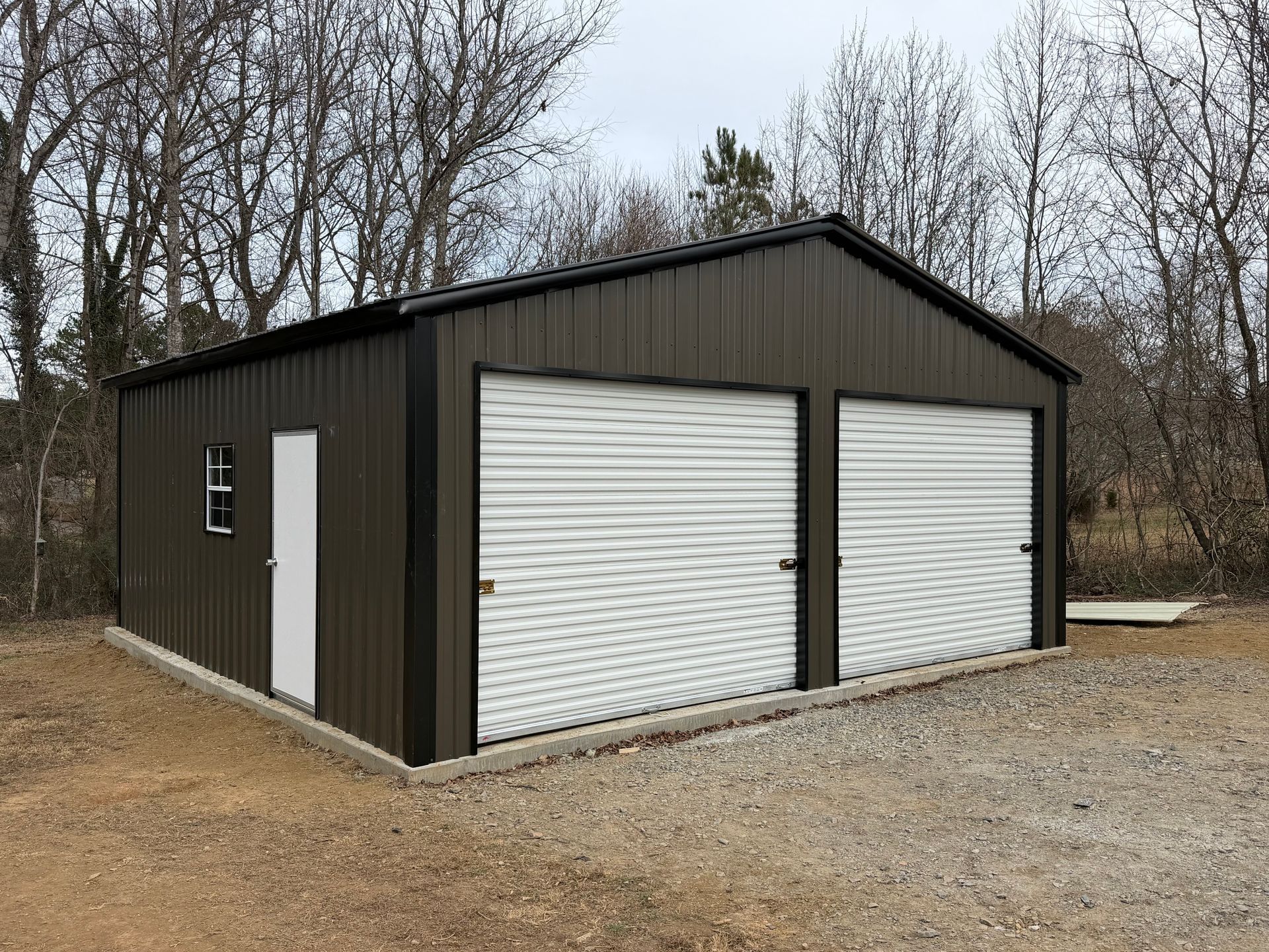 Dark metal garage with two white roll-up doors on a gravel lot beside bare trees.