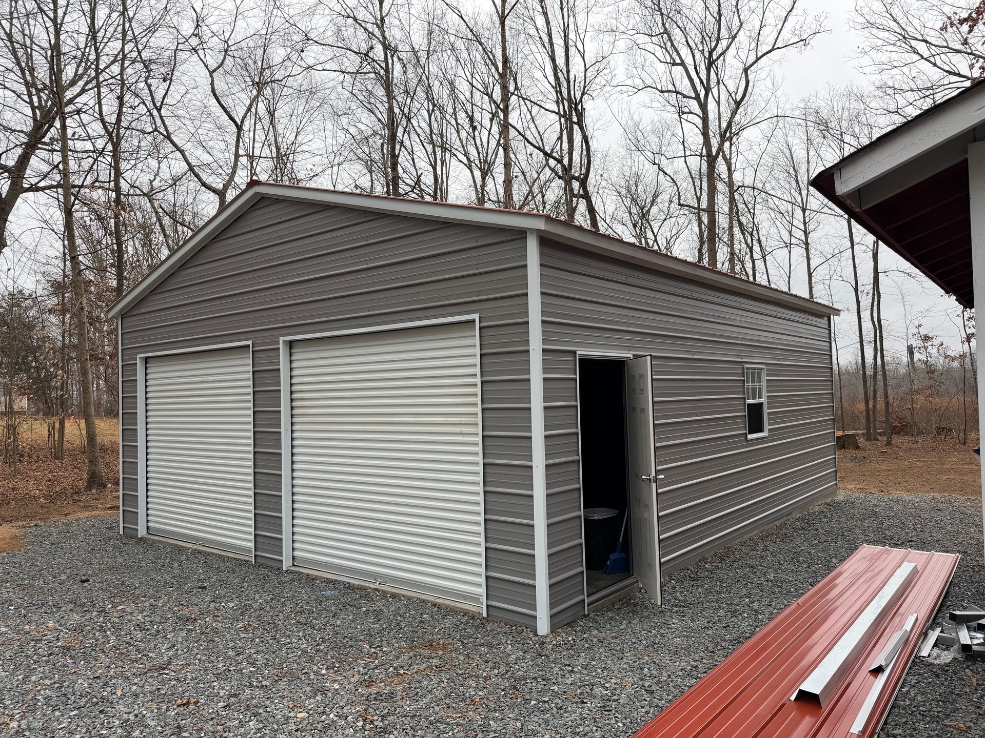 Gray metal shed with two roll-up doors and a side entrance on a gravel lot in a wooded area