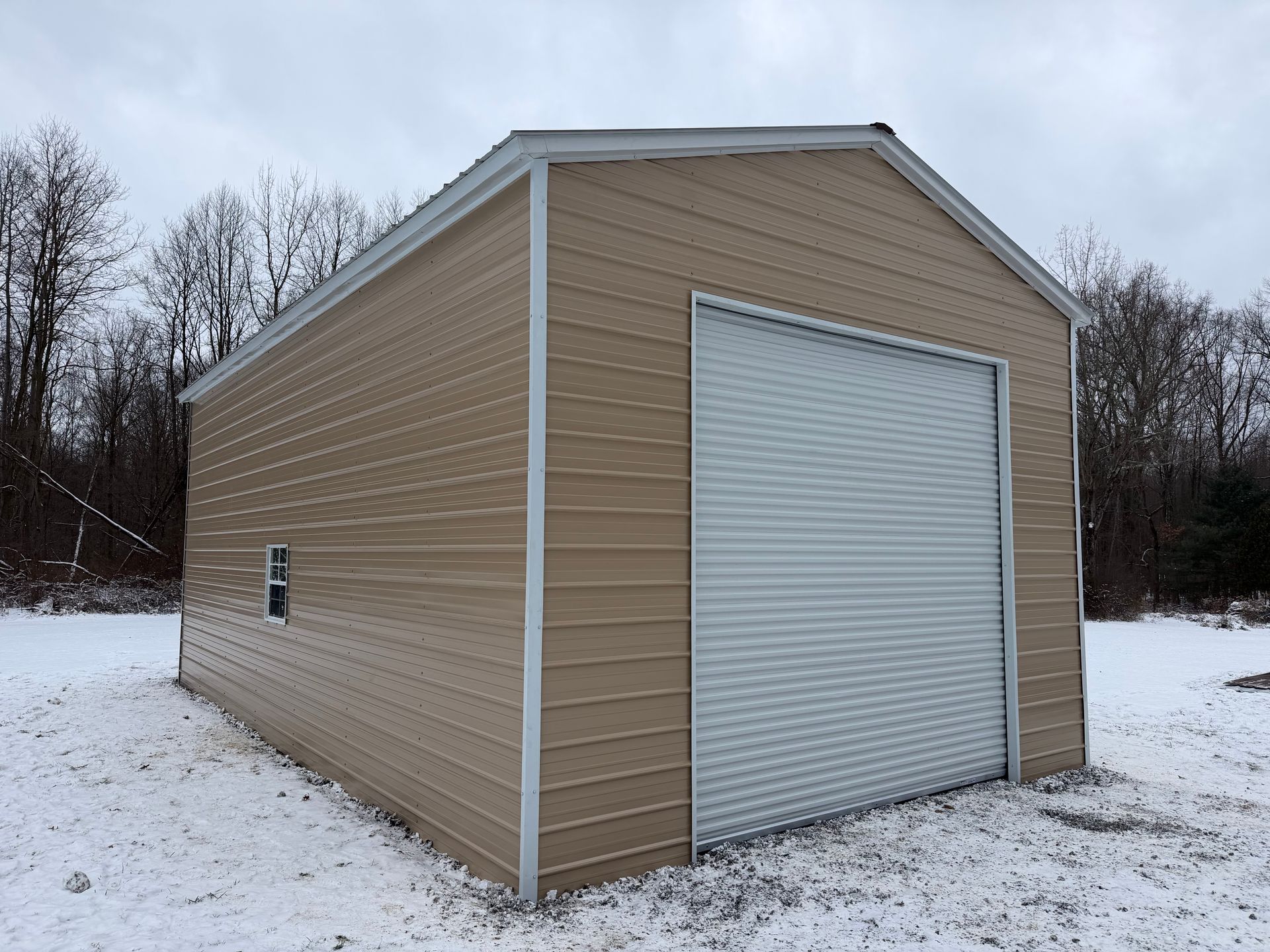 Tan metal-sided storage shed with a white roll-up door on a snowy lot