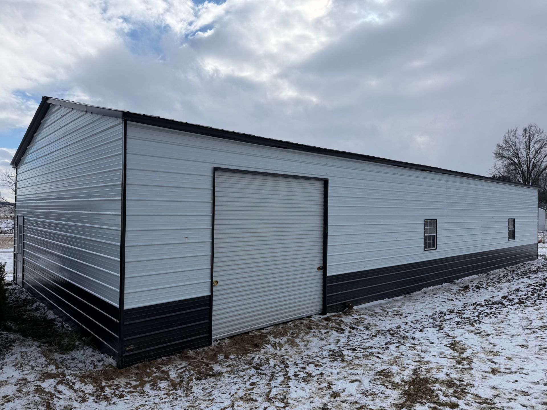 White metal building with a large garage door on a snowy lot under cloudy skies