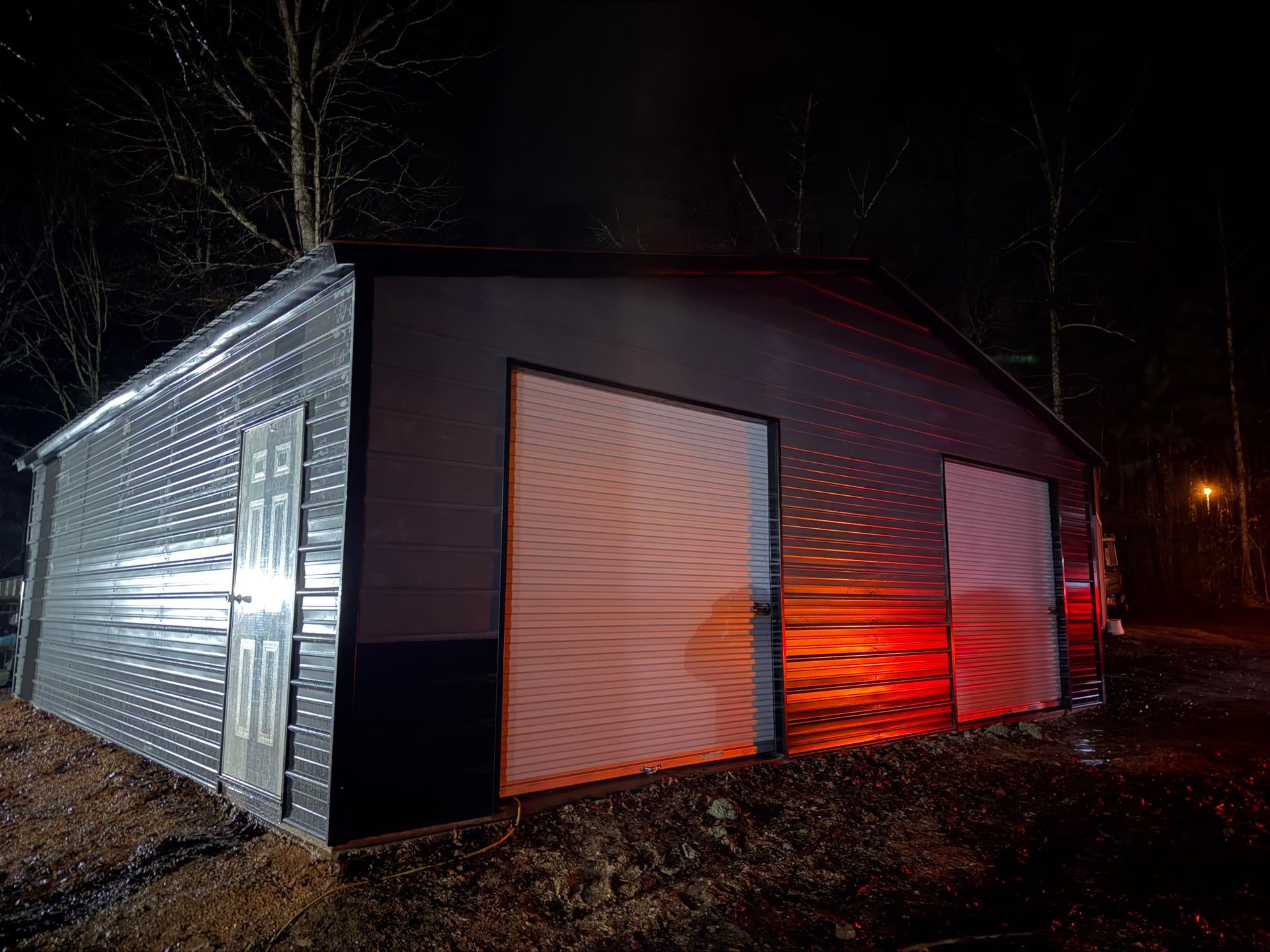 Small dark building at night with red light glowing through windows, surrounded by trees and dirt ground