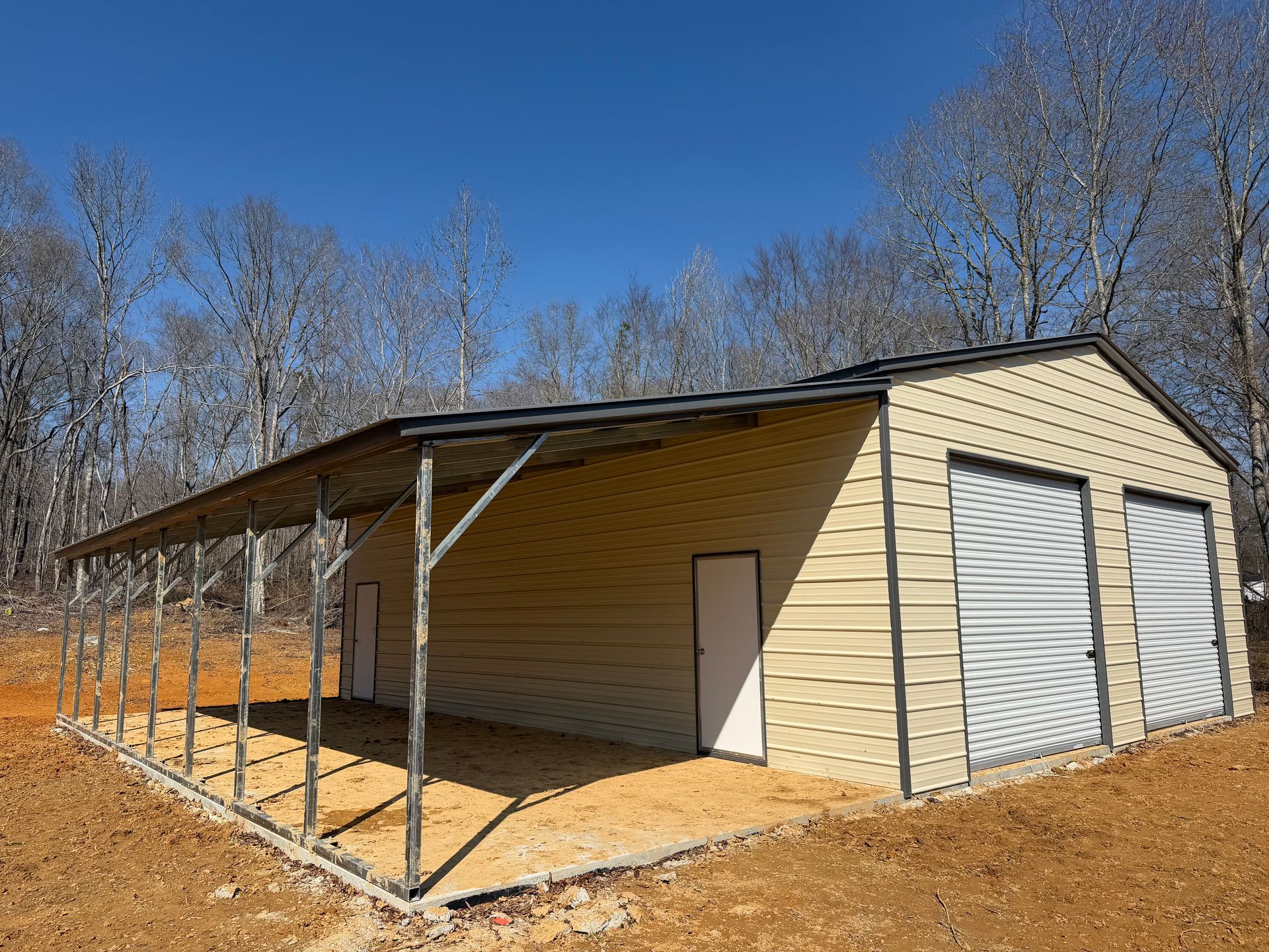Open-sided metal carport beside a beige garage in a wooded, leafless setting