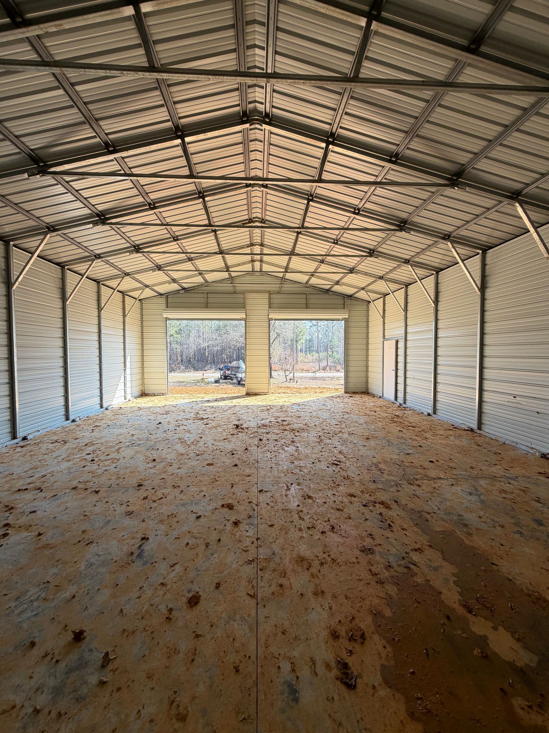Empty metal storage shed with open front, exposed beams, and dirt floor lit by daylight