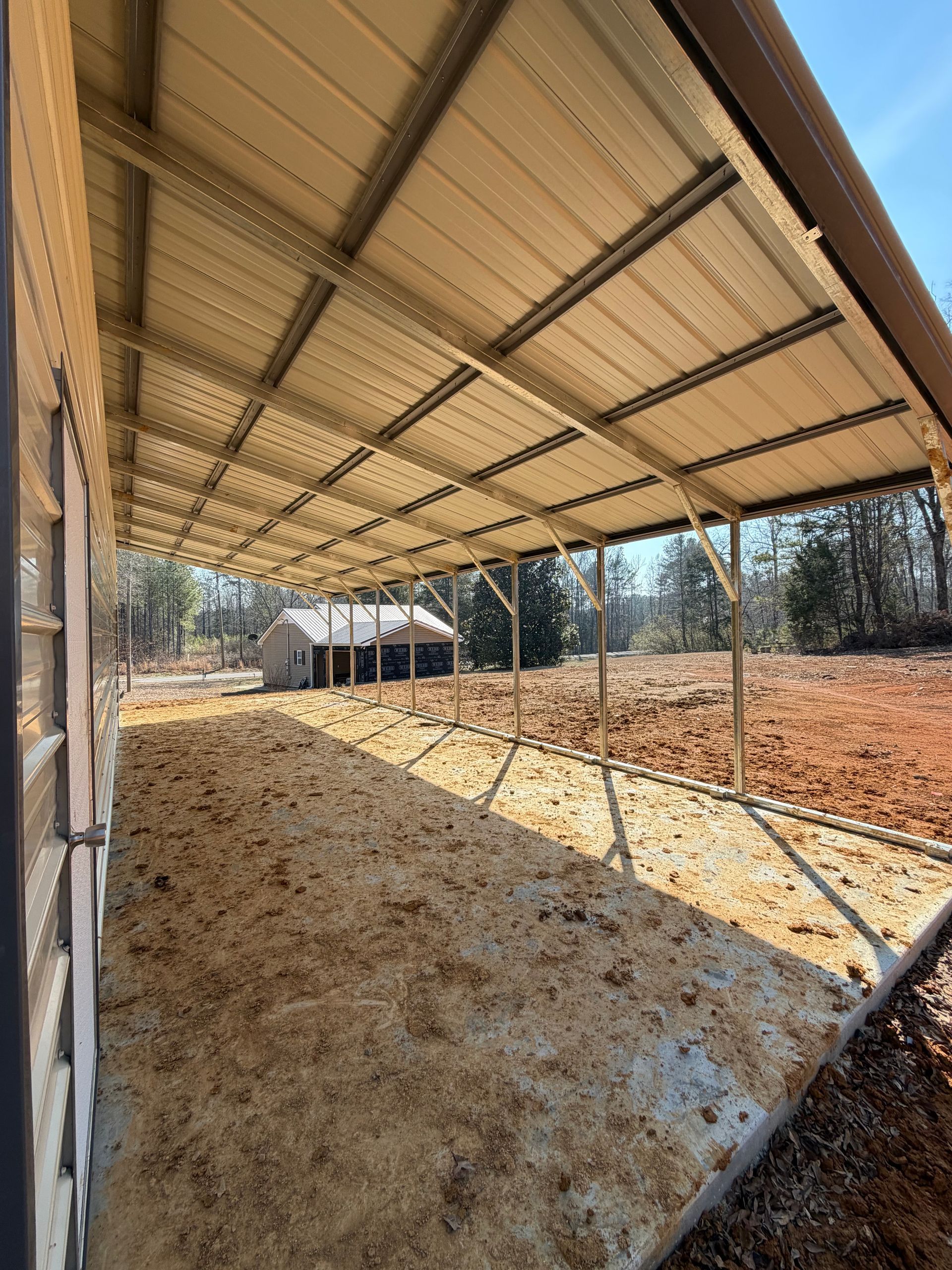 Covered livestock pen with feed under a metal roof, fenced with posts and wire in a rural area.