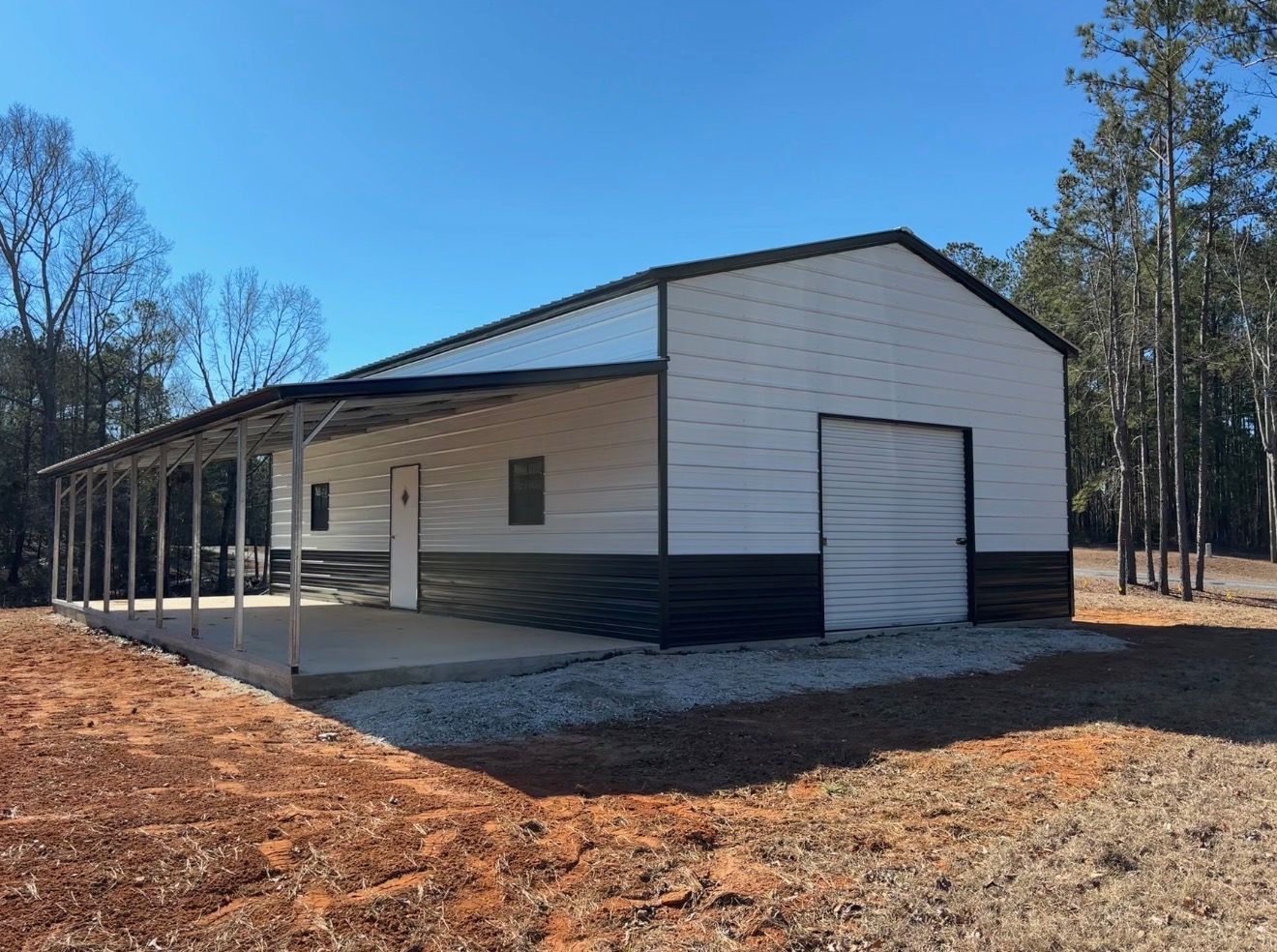 White metal barn with black trim and a covered porch in a wooded clearing under a clear blue sky