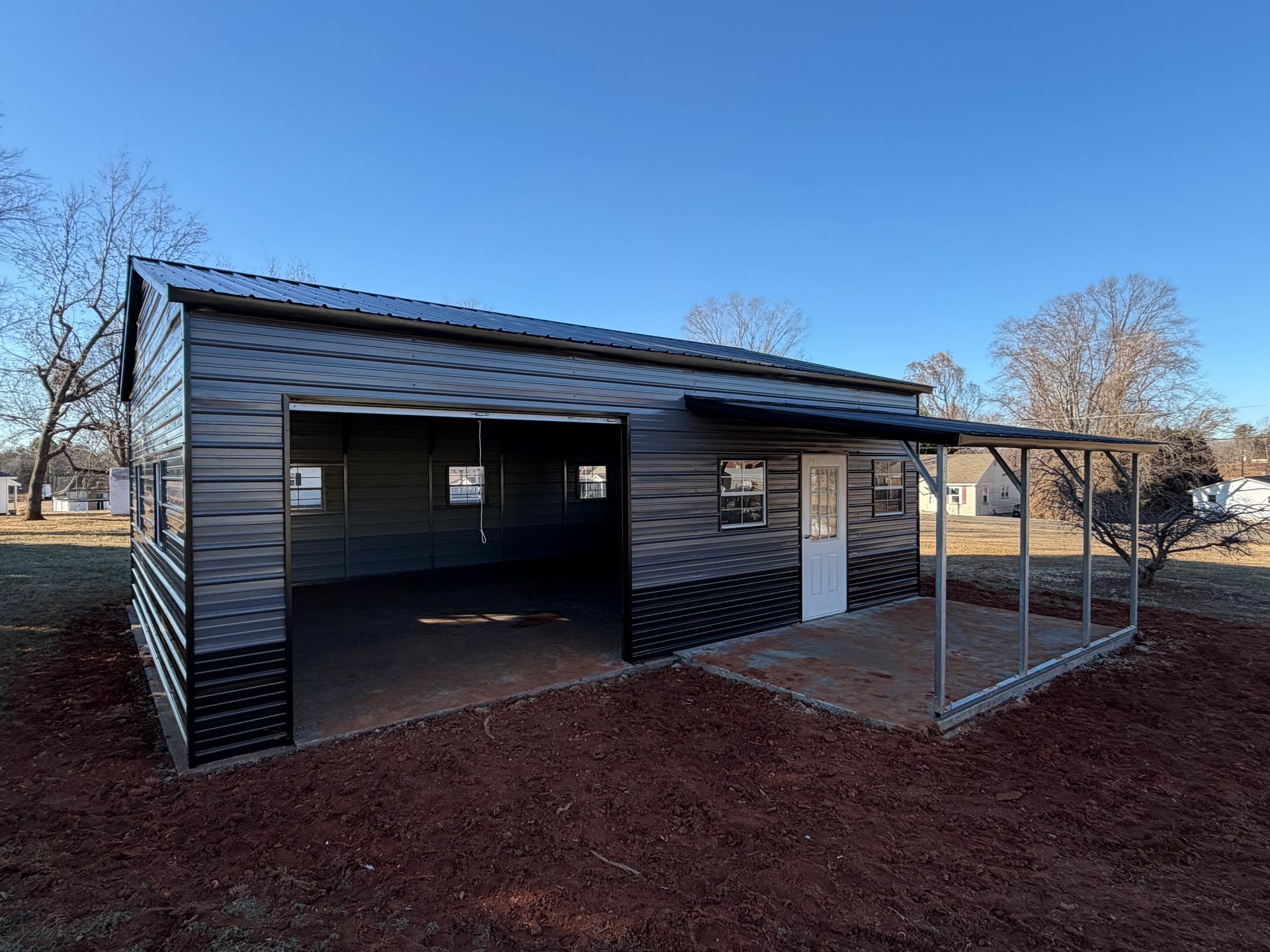 Small dark shed-like structure with open sides and a white door on a gravel lot under a blue sky