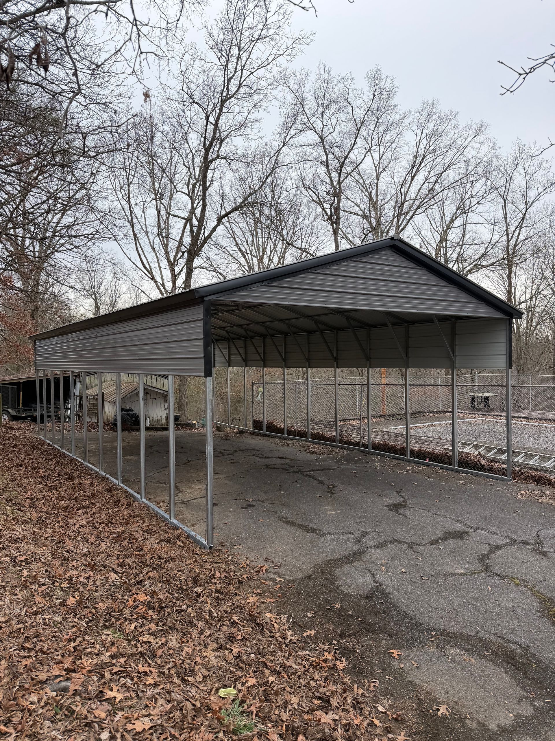 Empty metal carport beside a gravel lane in a leaf-covered, wooded area under bare trees