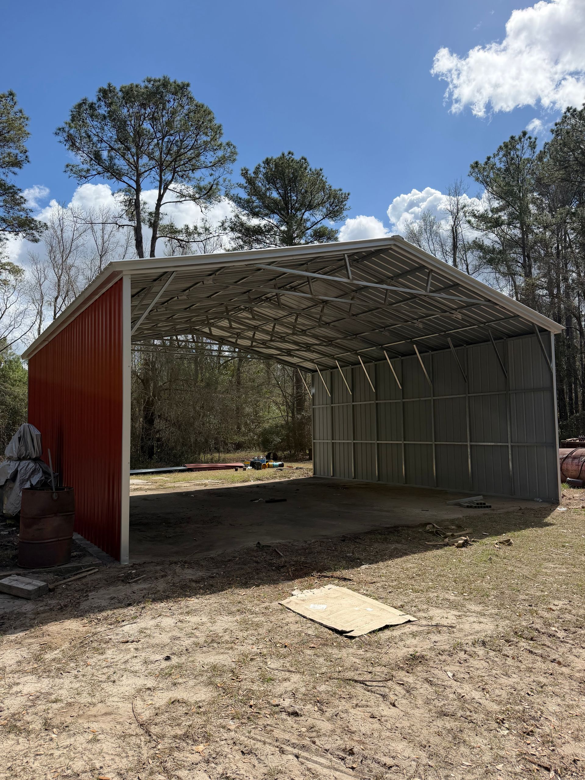 Open-sided red and gray shed frame on a dirt lot under a blue sky with trees in the background