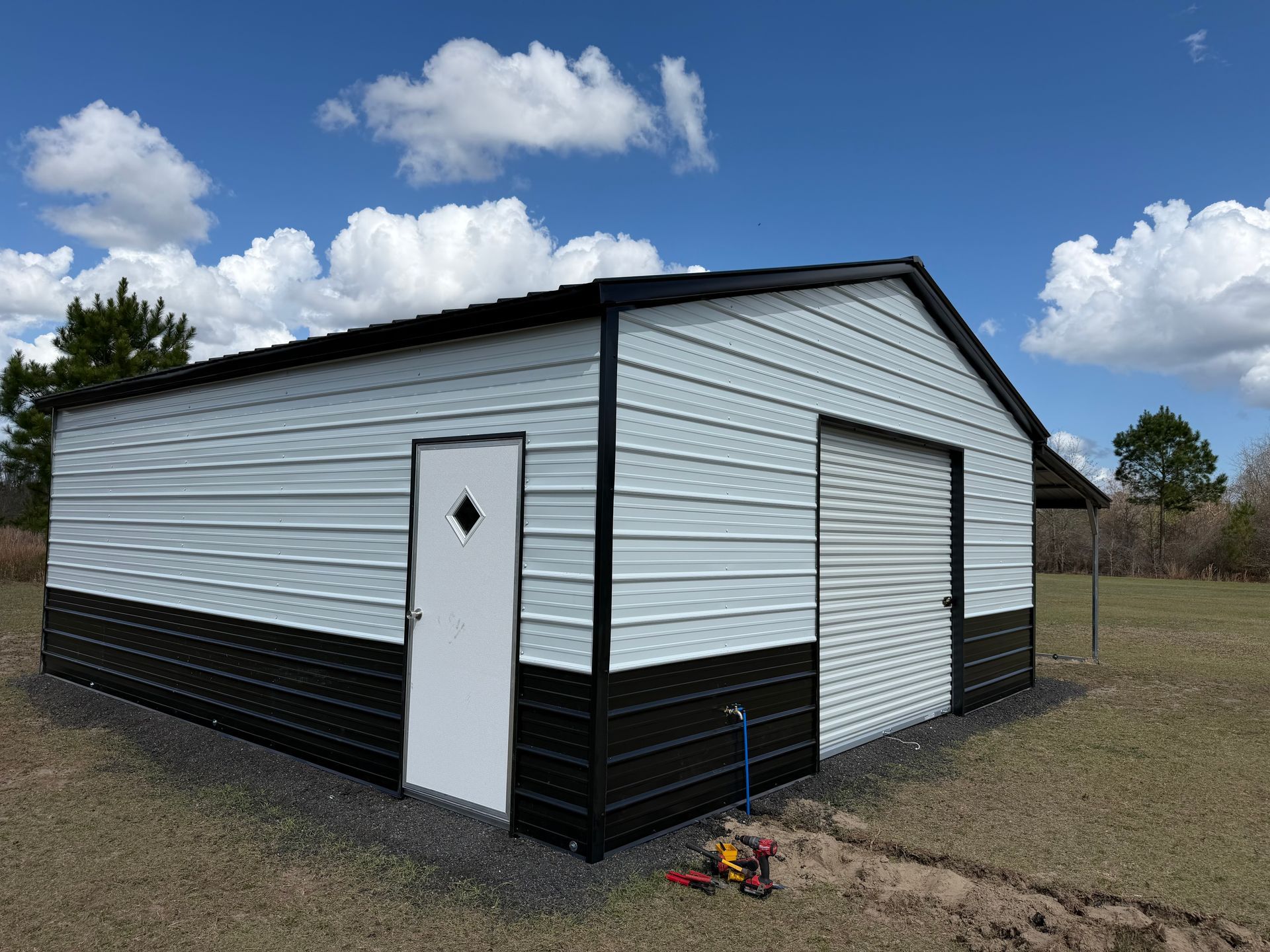 White-and-black detached garage with side door and large roll-up door on a gravel lot under a blue sky.