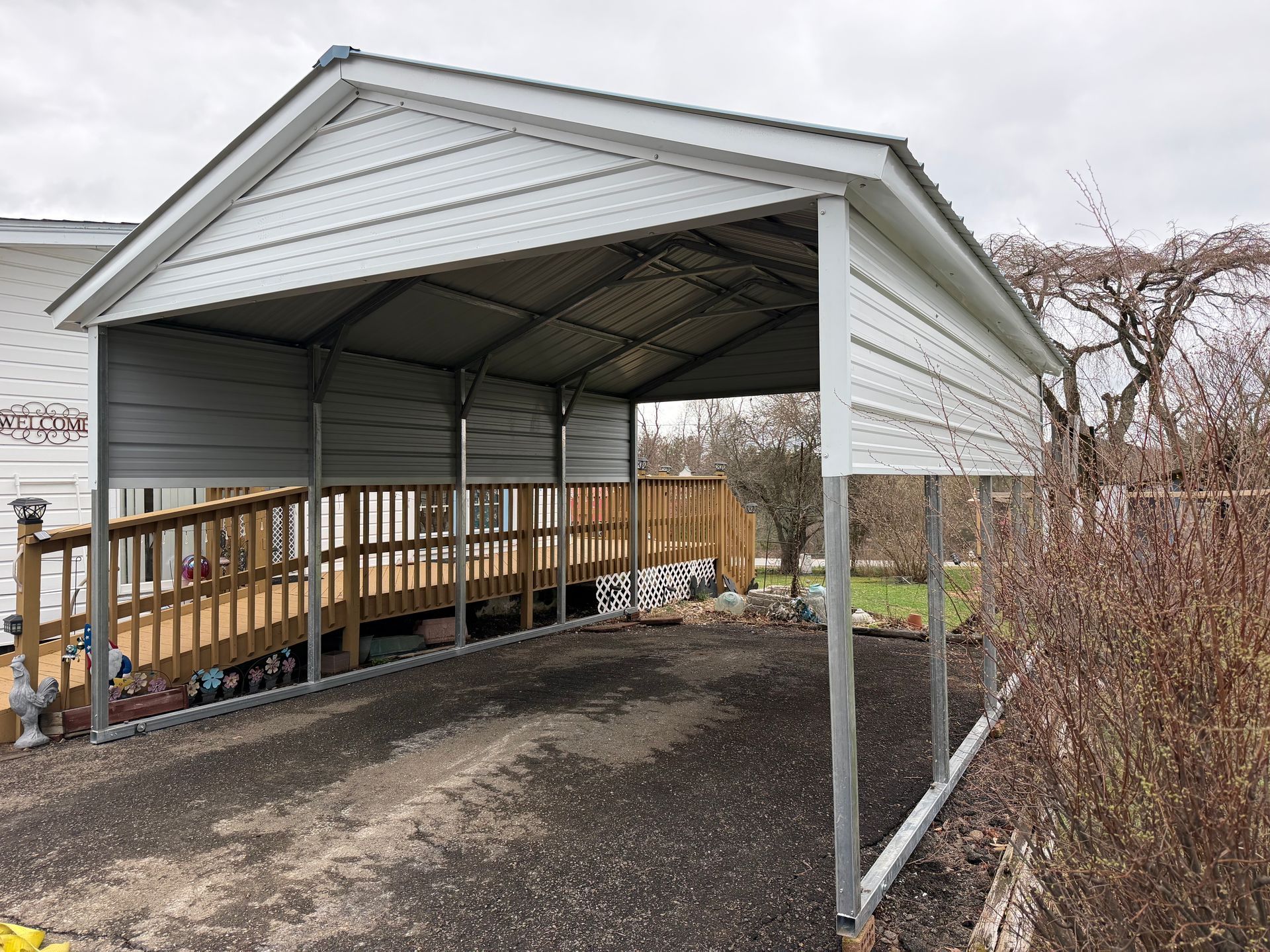 Gray metal carport attached to a house, covering a gravel driveway beside a wooden ramp.