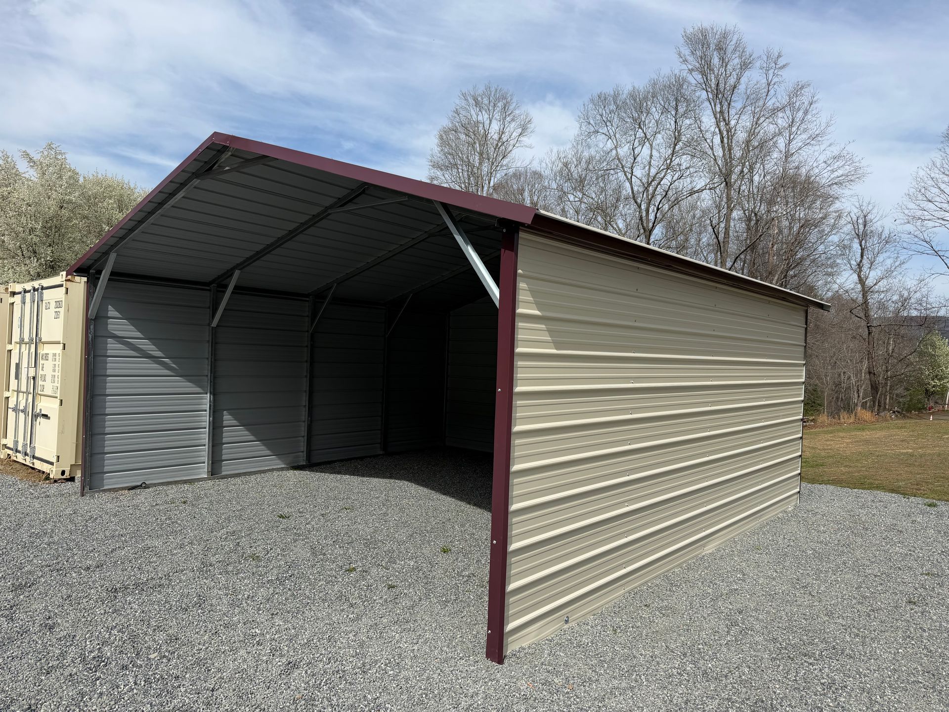 Metal carport shelter with beige siding and a gravel floor outdoors under a cloudy sky
