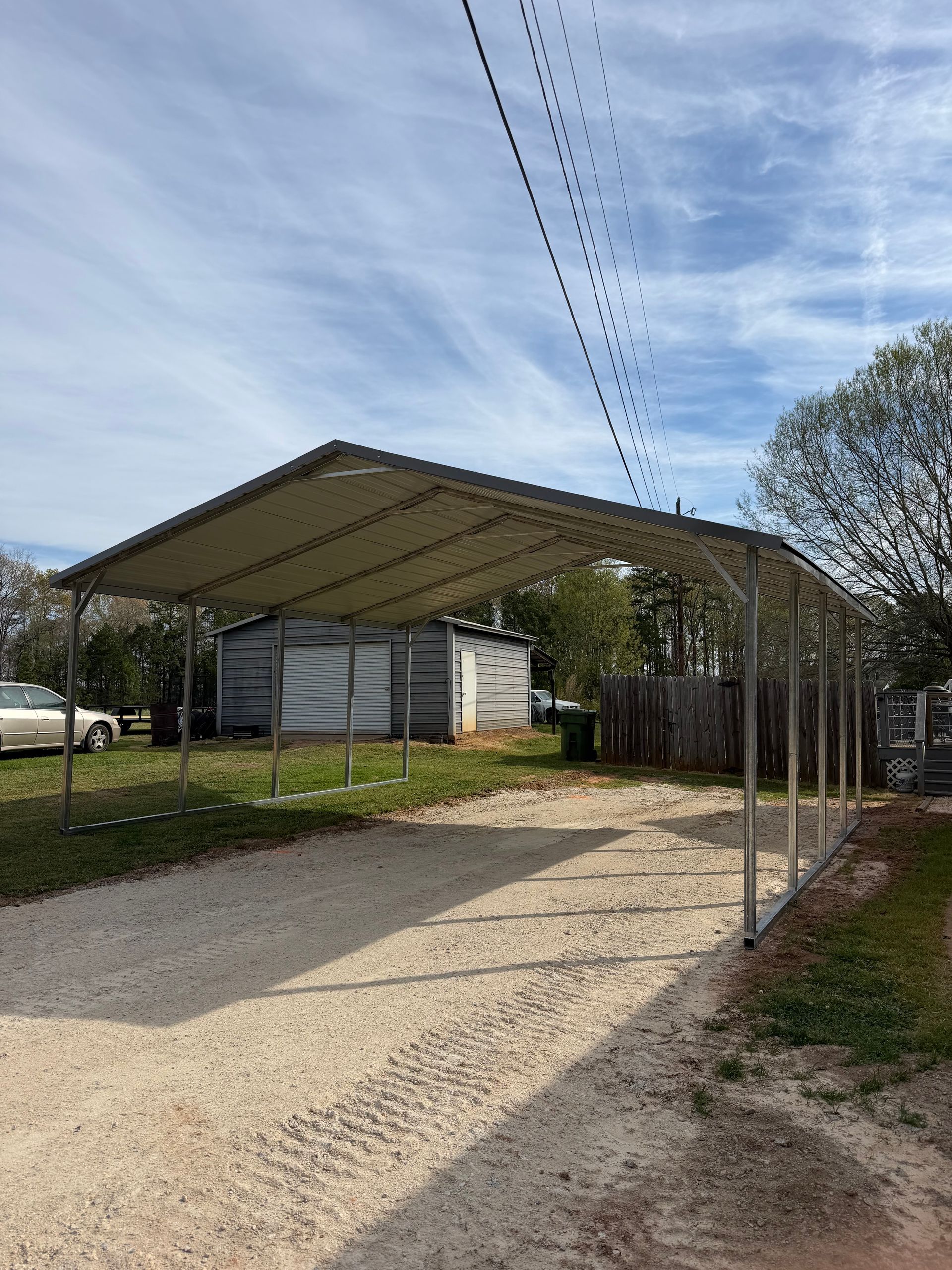 Gravel driveway leading to a large open metal carport beside a fenced yard under a cloudy sky