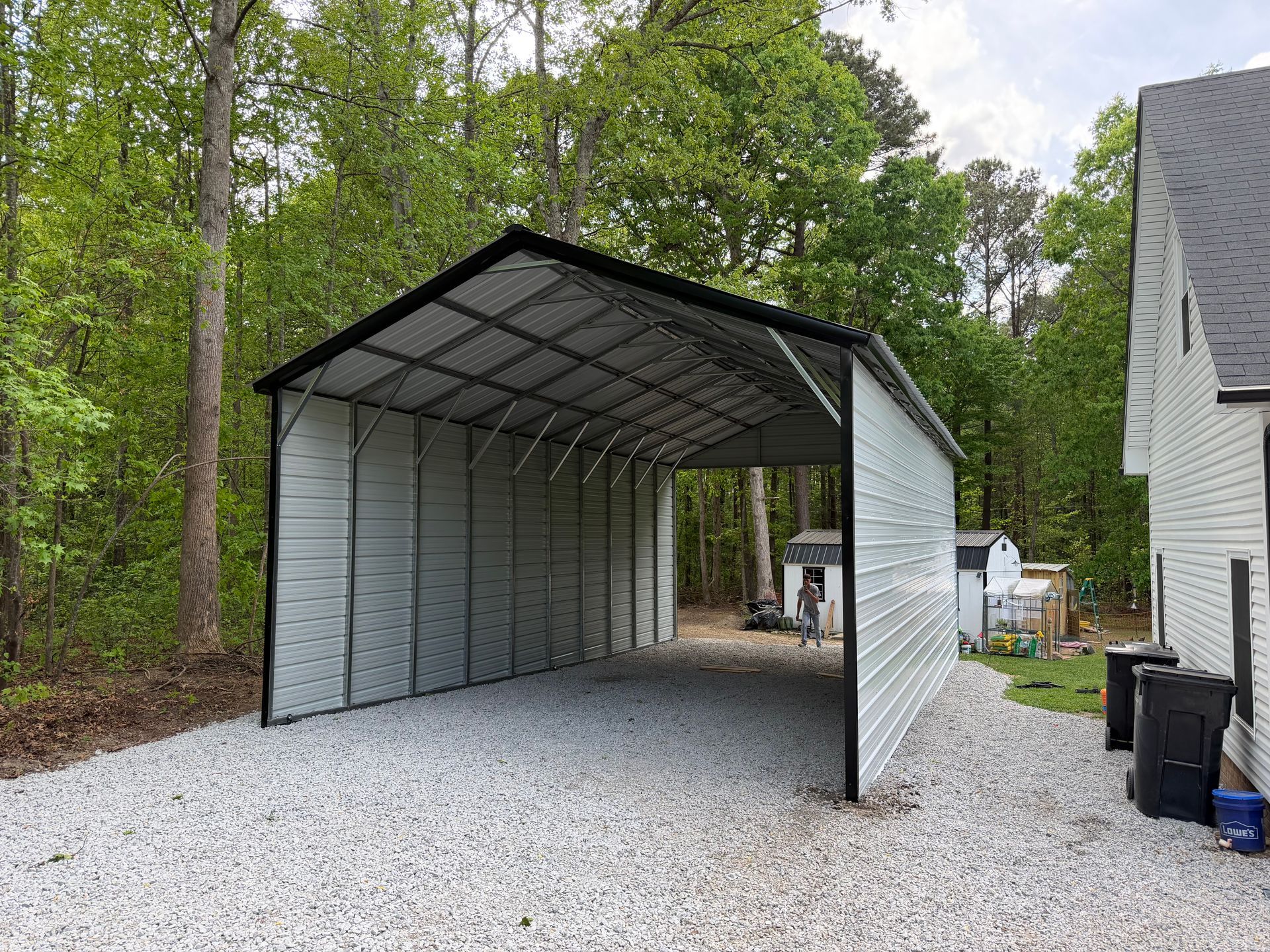 Open-sided metal carport on a gravel driveway beside a house and trees