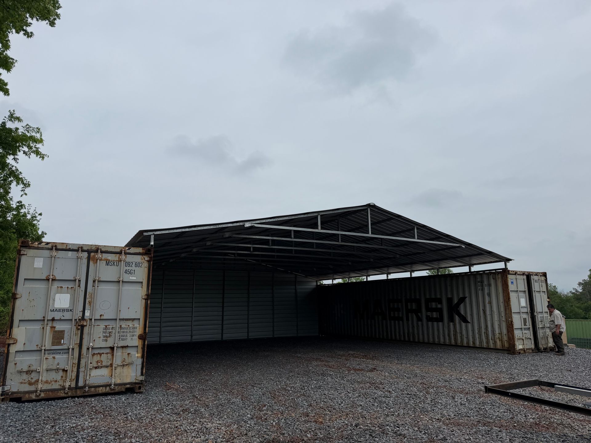 Collapsed warehouse roof with stacked shipping containers on both sides under a gray sky