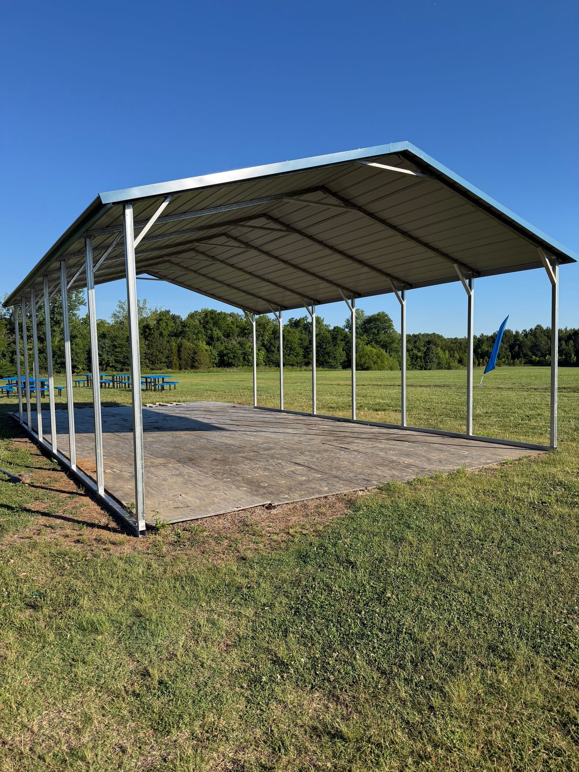 Open-sided metal-roofed pavilion on a gravel pad in a grassy field under clear blue sky