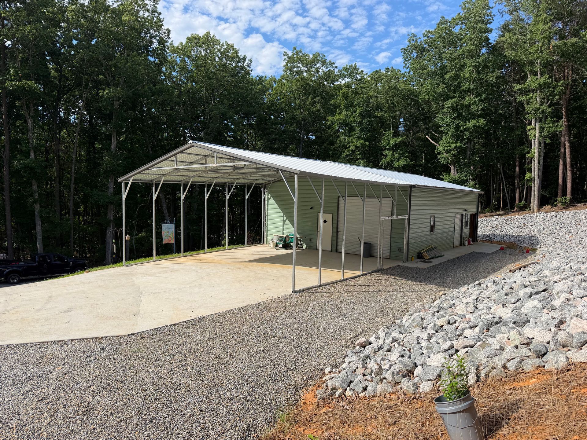 Open-sided metal carport on a gravel lot beside a wooded hillside under a blue sky