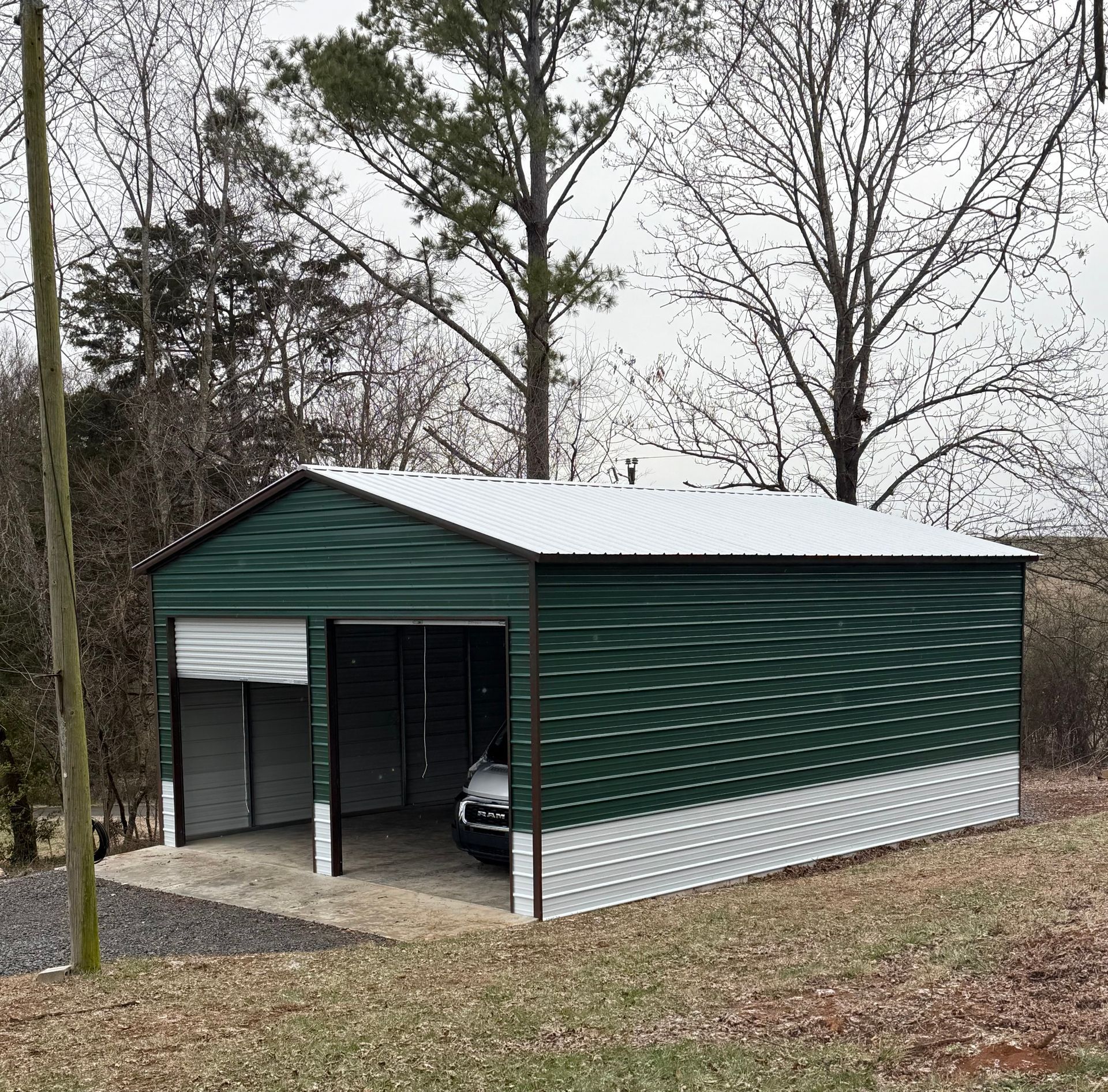 Green metal carport/shed with white roof and trim beside trees on a leaf-covered lot