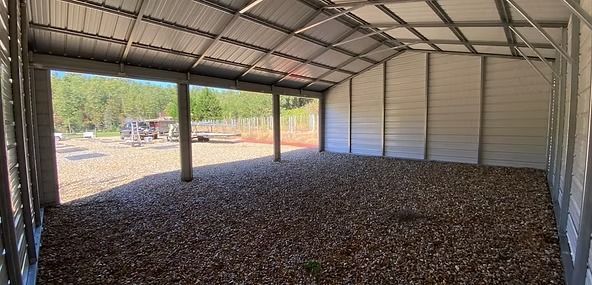Empty metal barn interior with open sides overlooking a gravel lot and trees outside