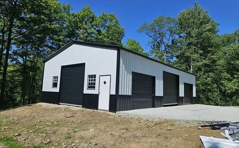 White metal garage with black doors on a concrete pad, set among trees under a clear blue sky.