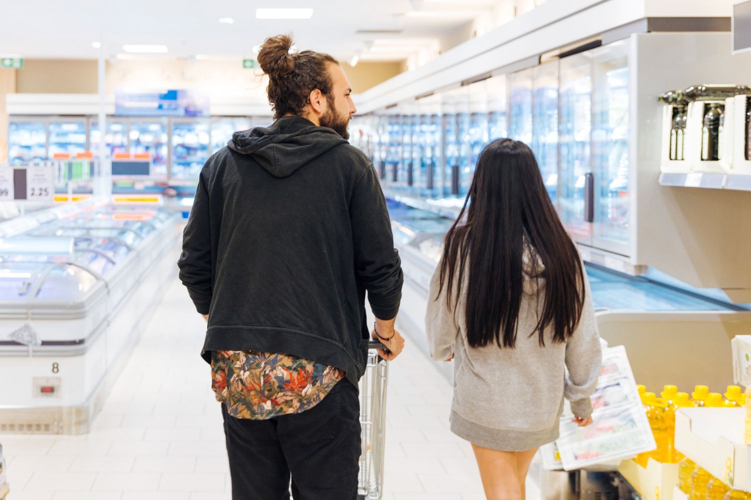 Couple walking down supermarket aisle, one pushing cart, other carrying items.