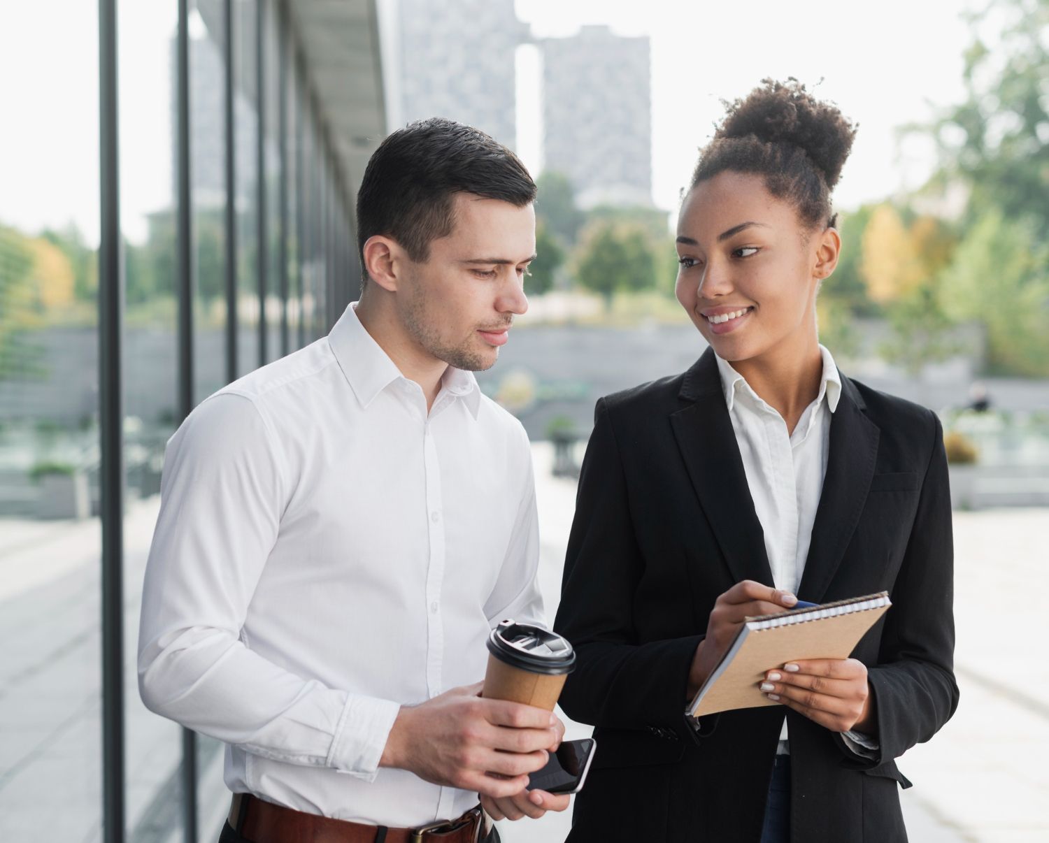 Man and woman in business attire converse outside a building. The woman holds a notepad.