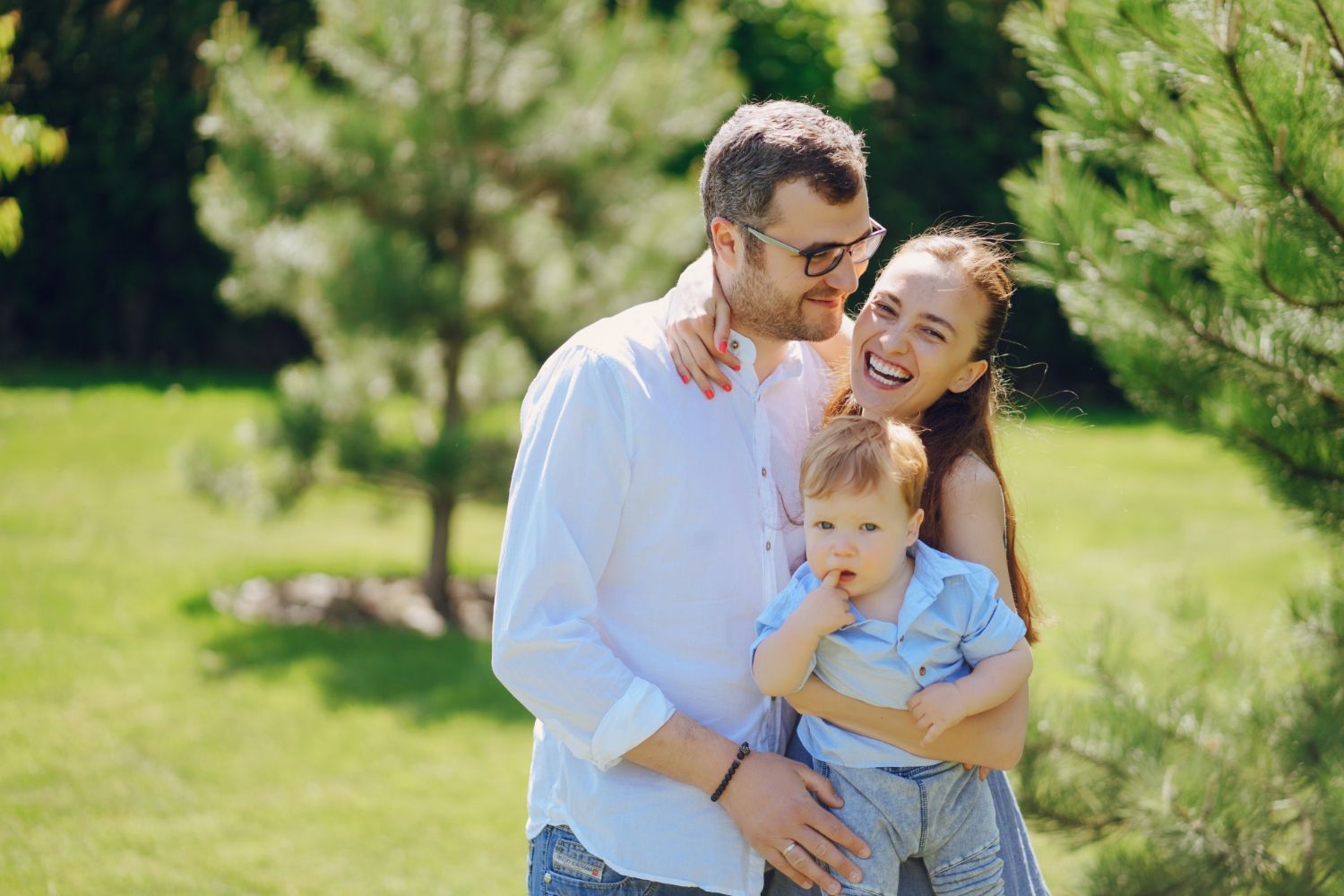 Family embracing outdoors: Man, woman, and toddler smiling on grassy lawn; green trees in background.