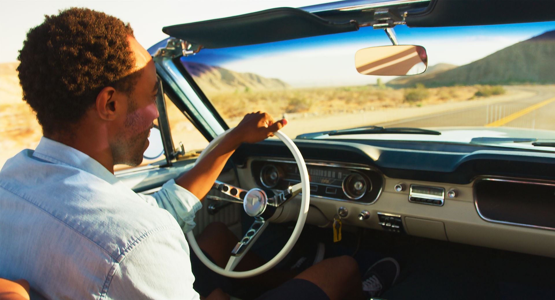Man driving a classic convertible on a desert road, sunny day.