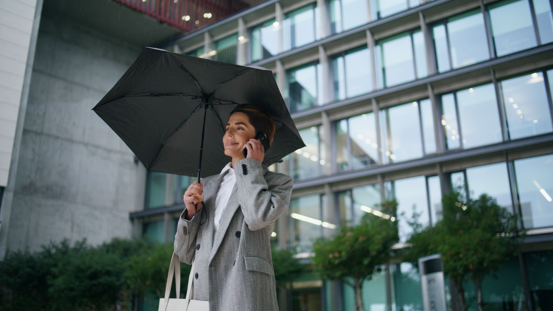 Woman in grey blazer holding umbrella, talking on phone outside modern building.
