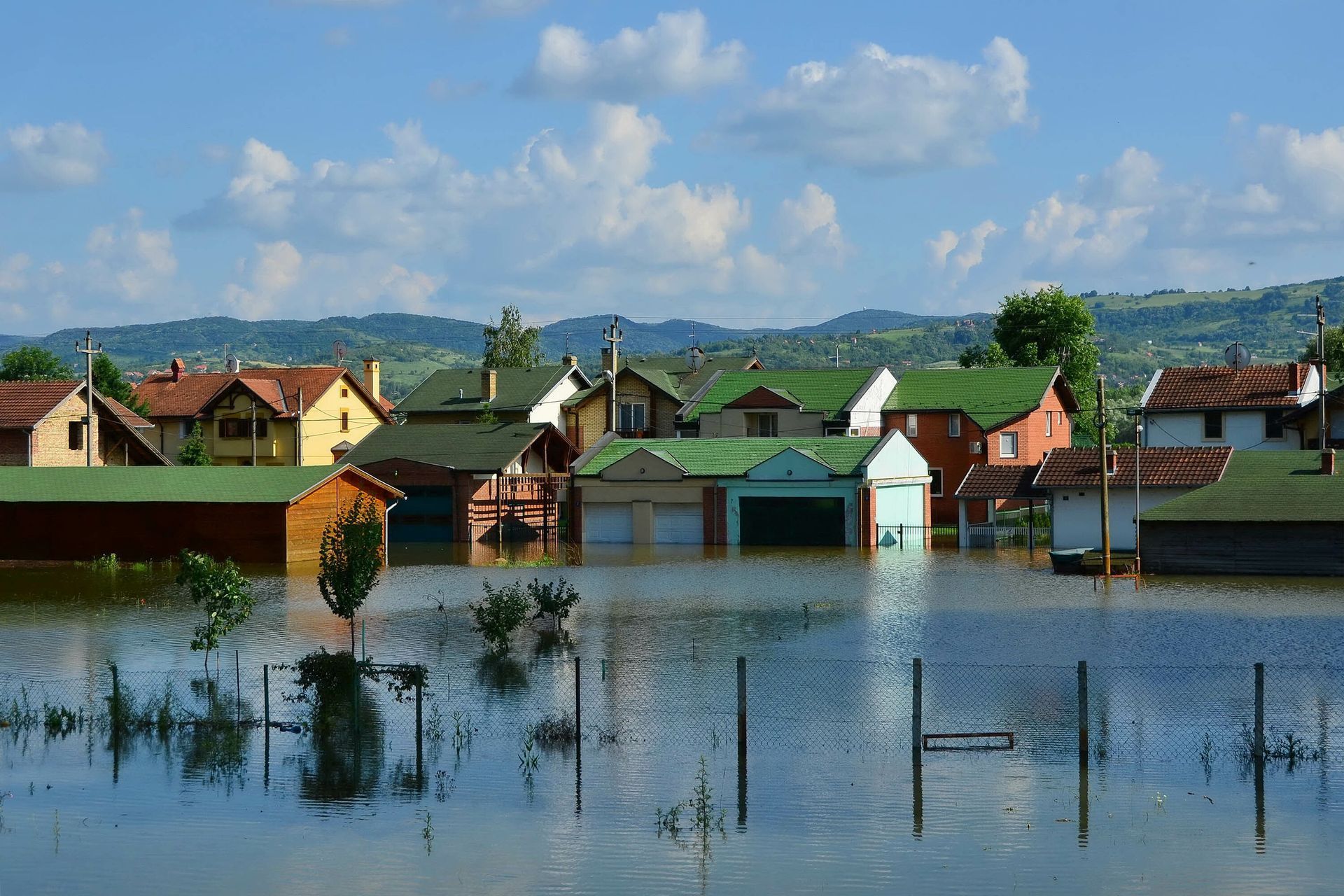 Flooded houses in a town; water covers the ground, trees visible, blue sky with mountains in the background.