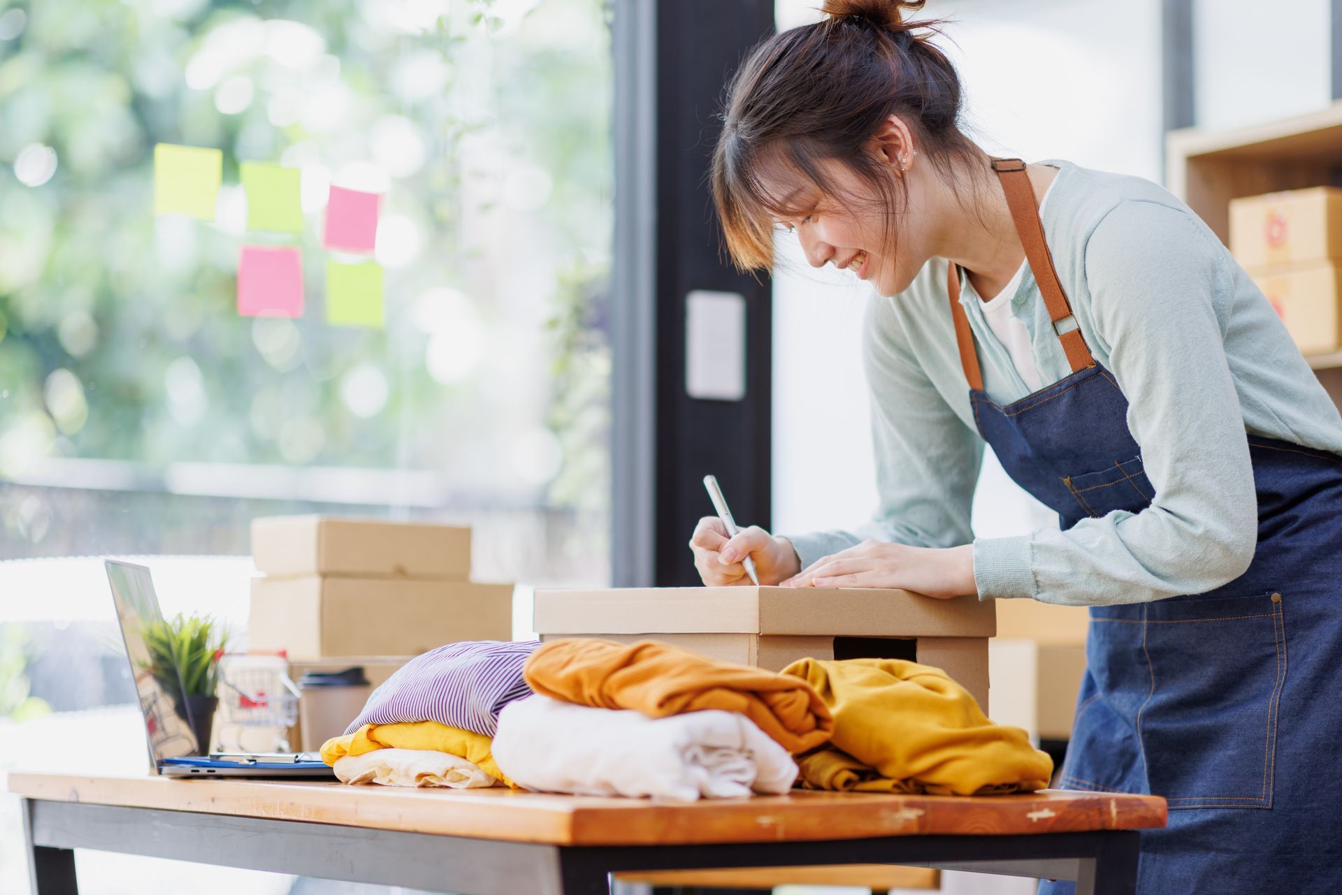 Woman in apron writing on a box; clothing, laptop, and packages on a table.