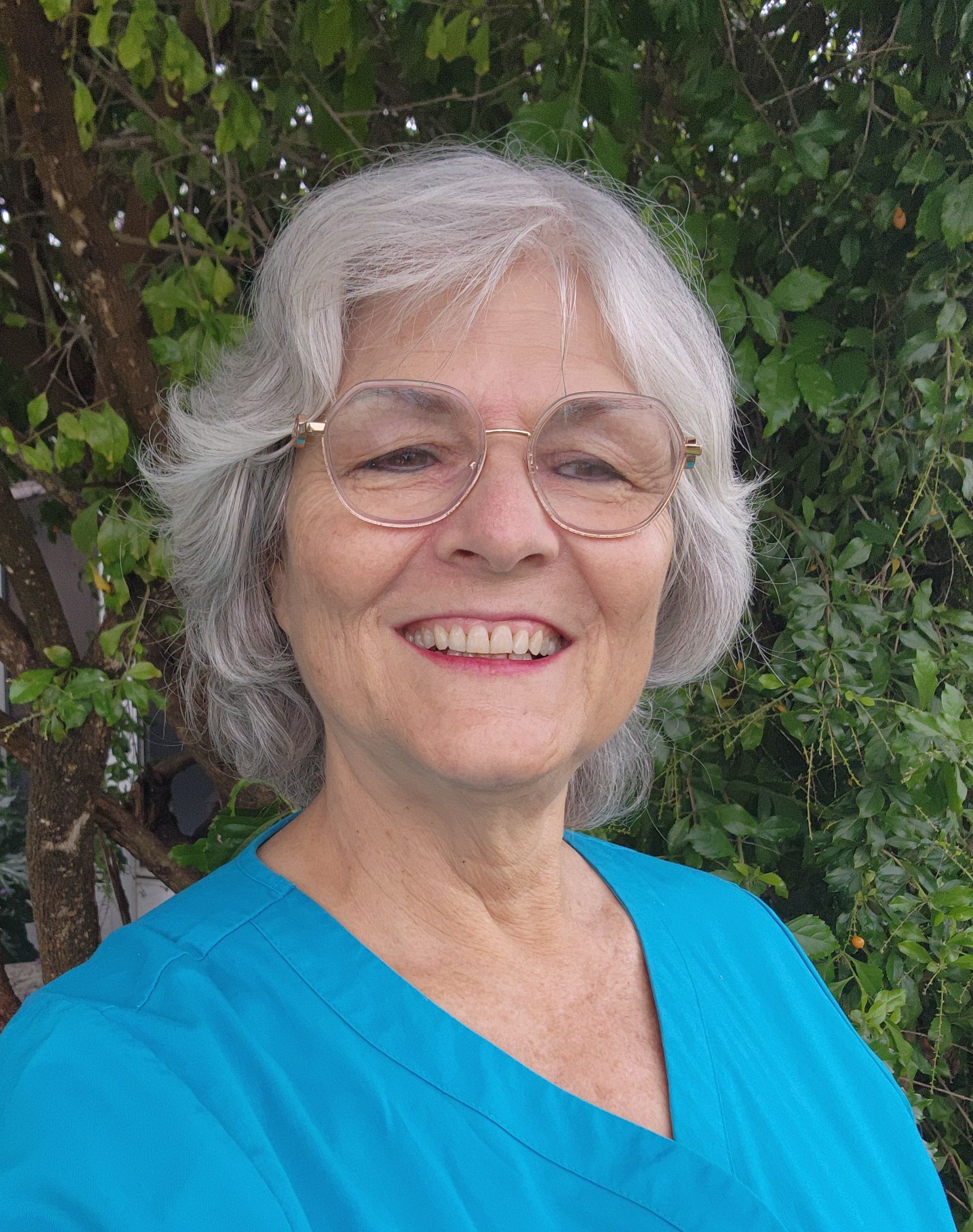 Woman with glasses, gray hair, smiling, wearing a blue shirt. Green foliage background.