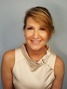 Woman with blonde hair, smiling, wearing a sleeveless cream-colored top, in front of a blue background.
