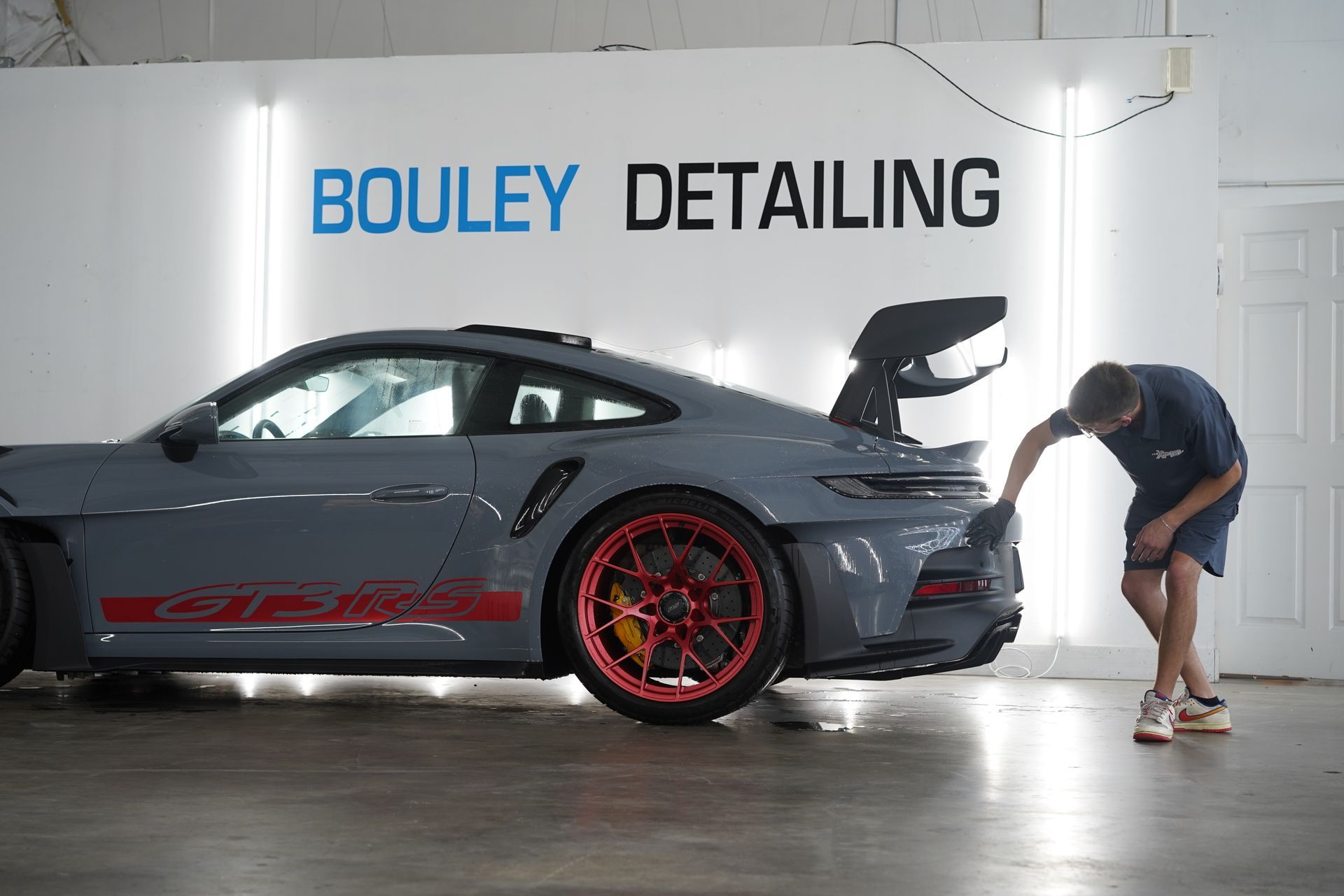 A man detailing a grey Porsche GT3RS with red accents in a shop. The shop has 